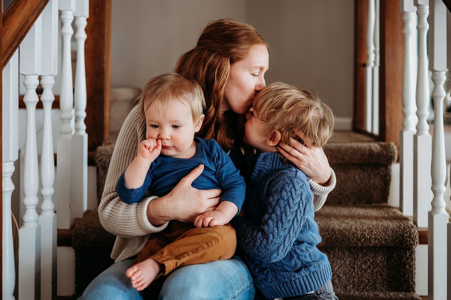 A mother sits on stairs hugging two young children, sharing a tender family moment during an Indianapolis in-home photo and video session.