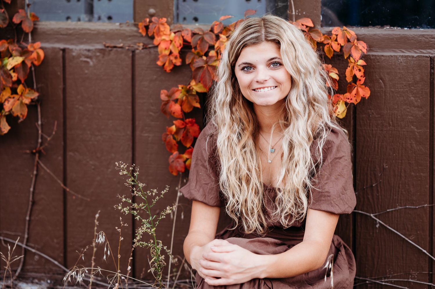 A person with long blonde wavy hair wearing a brown dress poses outdoors against a garage door with orange autumn vines.