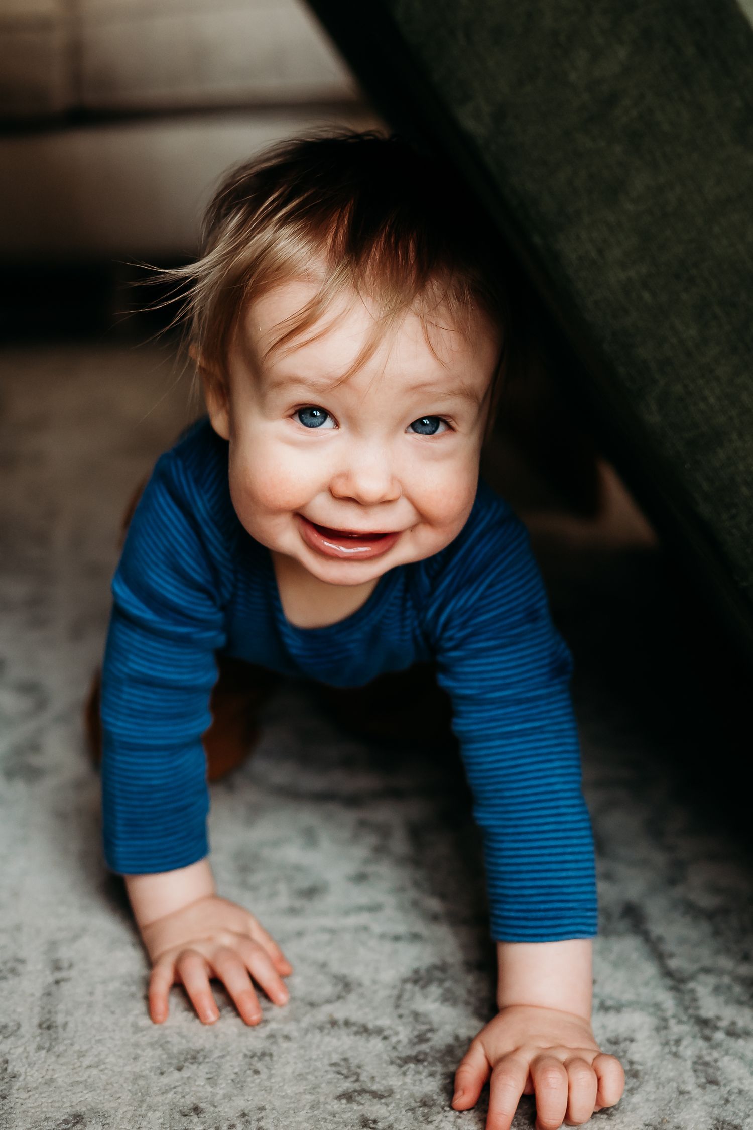 Smiling baby in blue striped onesie crawling on gray carpet near a couch, looking up with bright blue eyes.