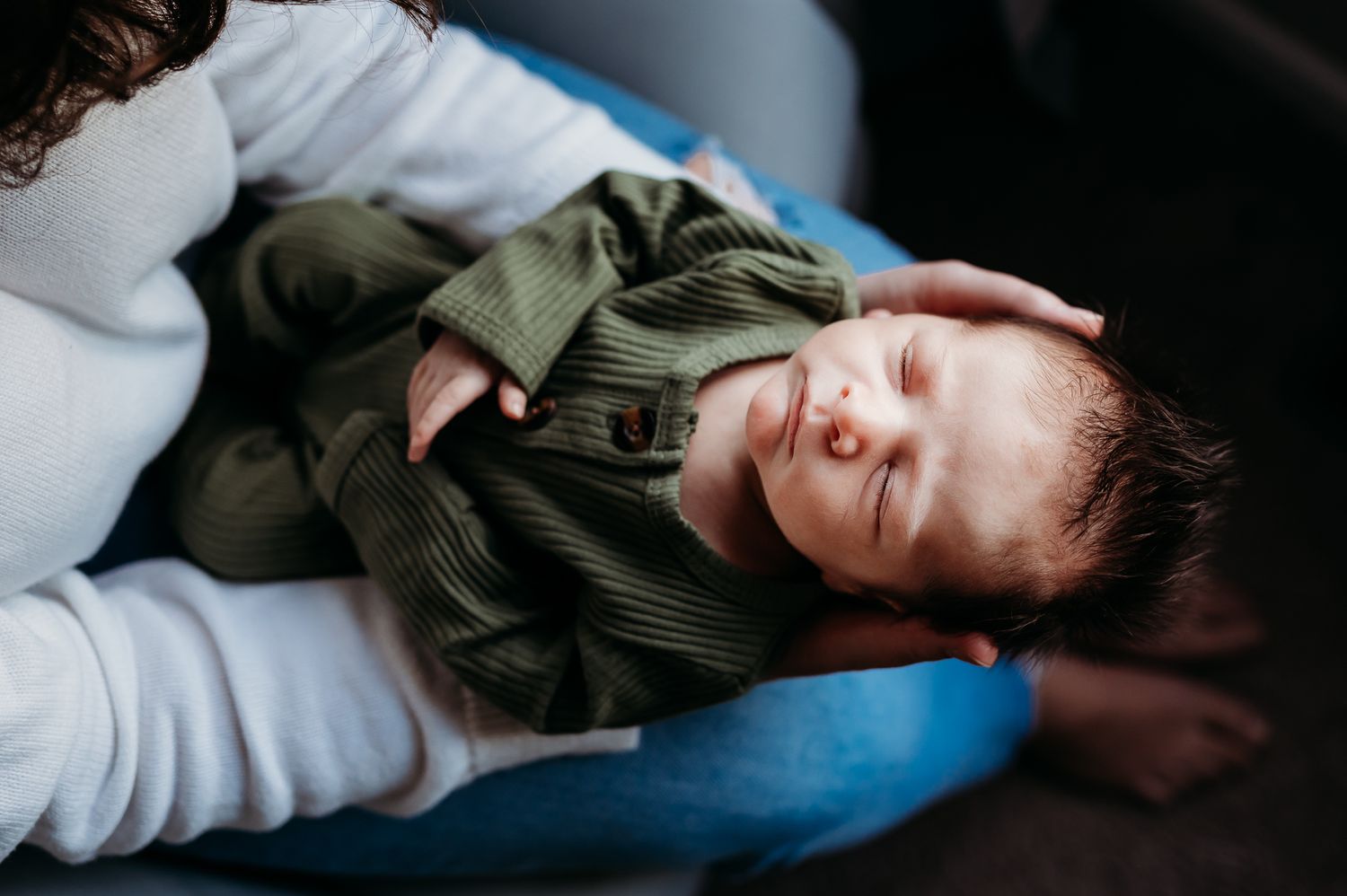 A baby wearing green clothing lies peacefully on mother's lap during an in-home photo session in Noblesville, Indiana.