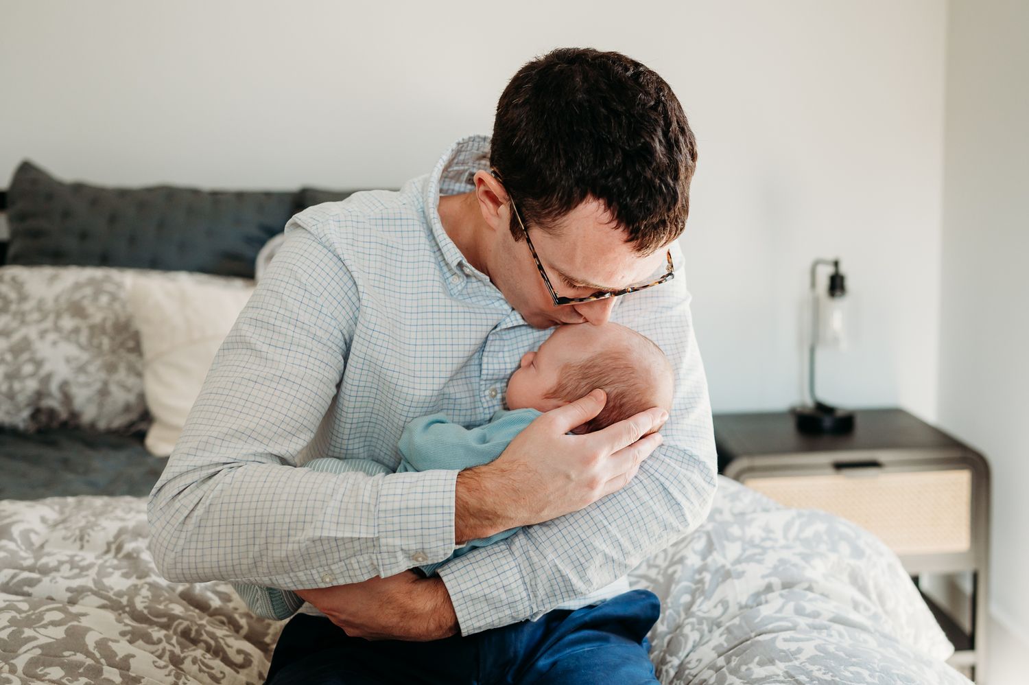 Westfield, Indiana, father in a light blue button-down shirt sitting on a bed tenderly holding a sleeping newborn baby.