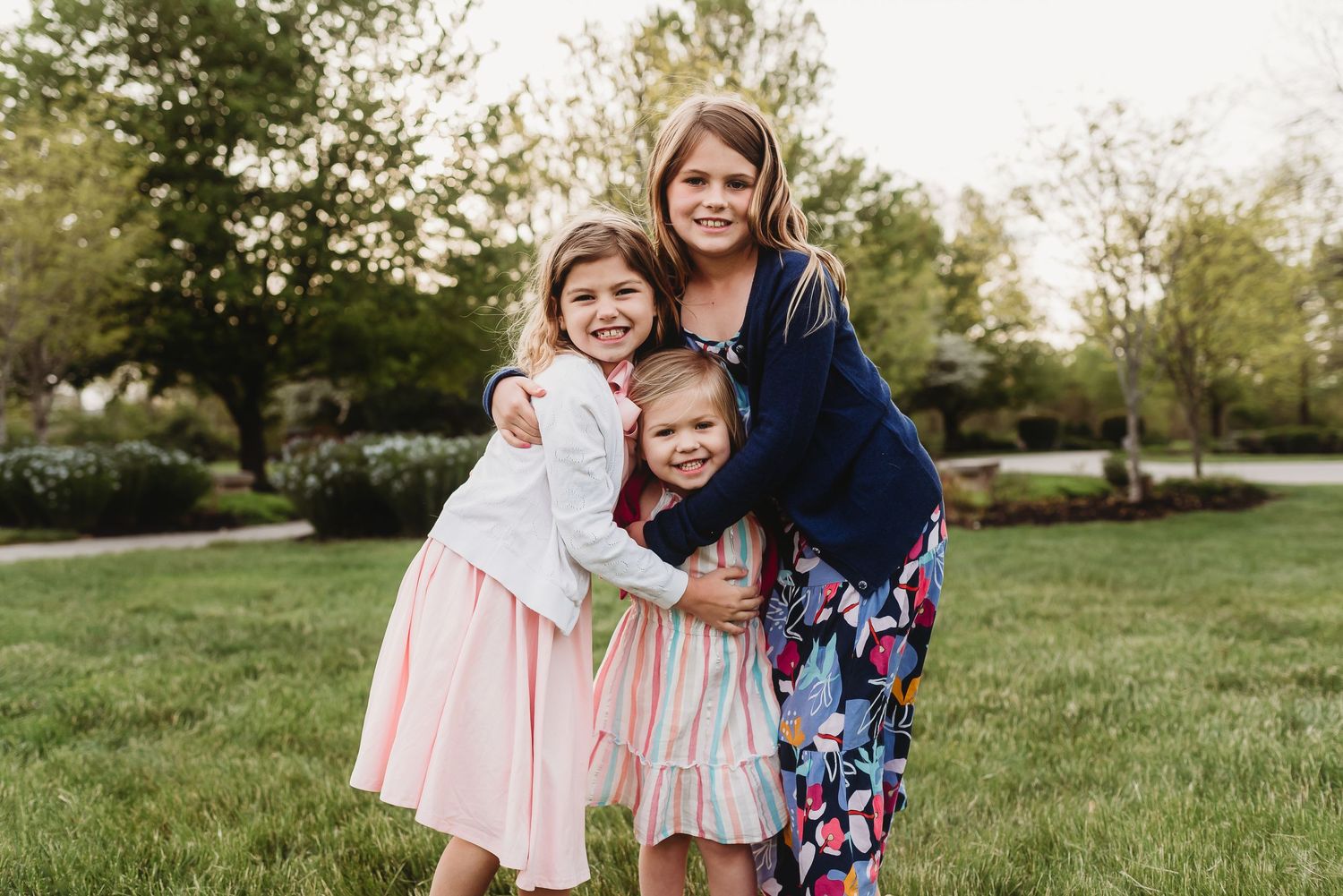 Three sisters in coordinating pink and blue outfits embrace while standing on green grass in a park setting in Plainfield, Indiana.