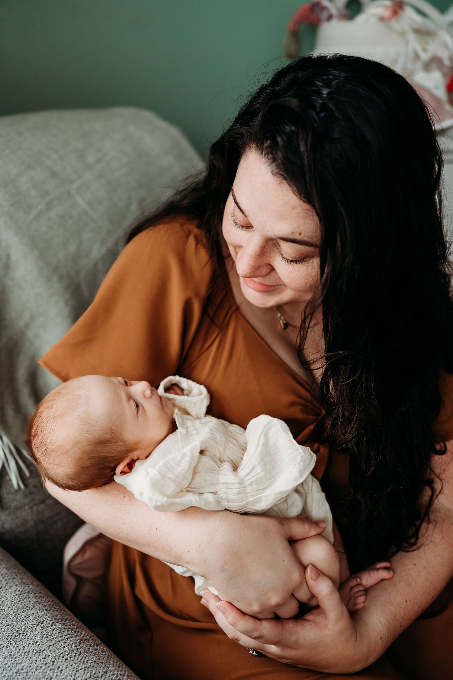 A tender moment captured as a Noblesville, Indiana, mom in a rust-colored shirt cradles a newborn baby in rocking chair in nursery.