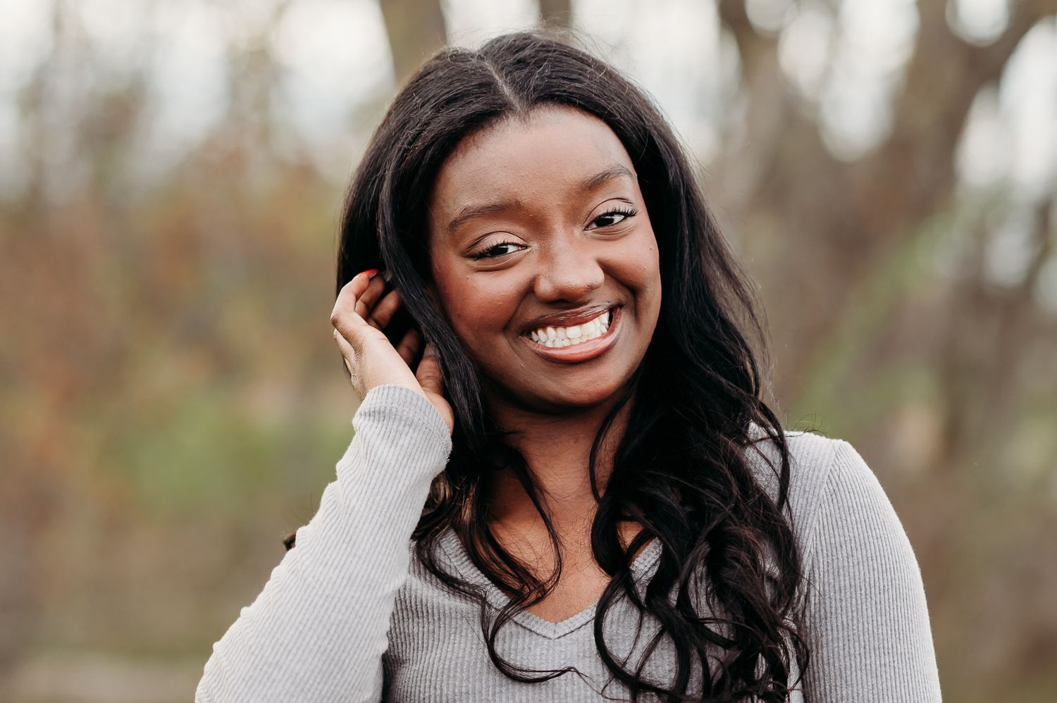 A radiant smile gleams as long dark hair cascades over a light gray sweater outdoors.