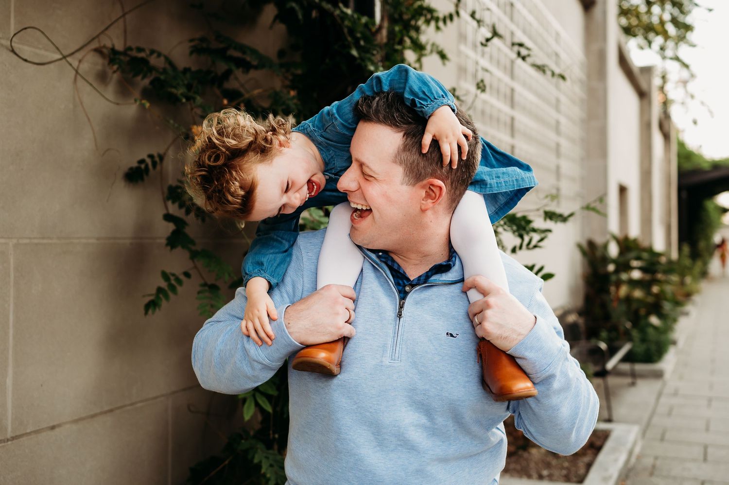 A playful moment captured outdoors as a child sits on shoulders near a light colored wall with climbing vines.