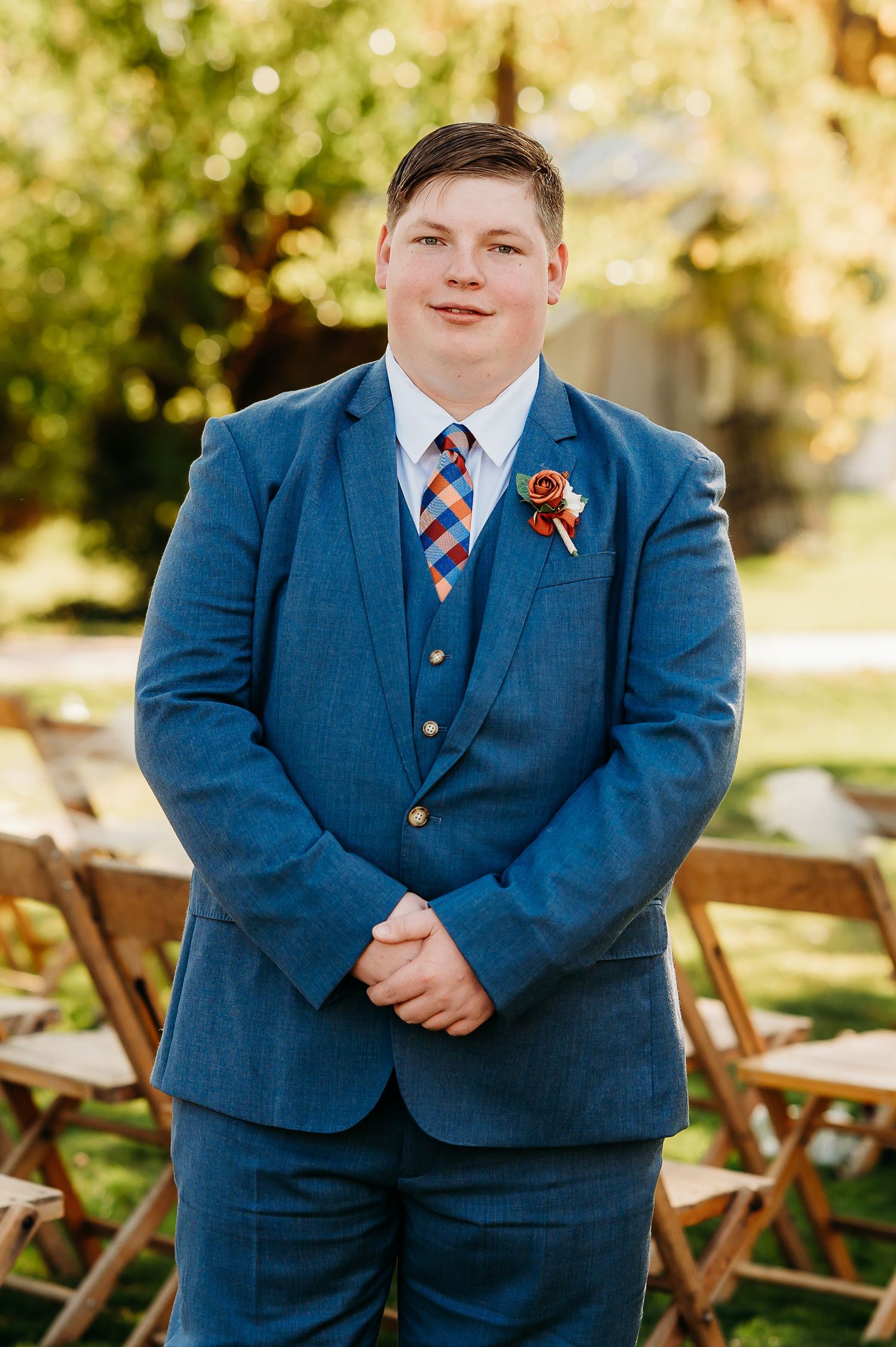 Person in navy blue suit with striped tie poses outdoors at wedding venue with wooden folding chairs.
