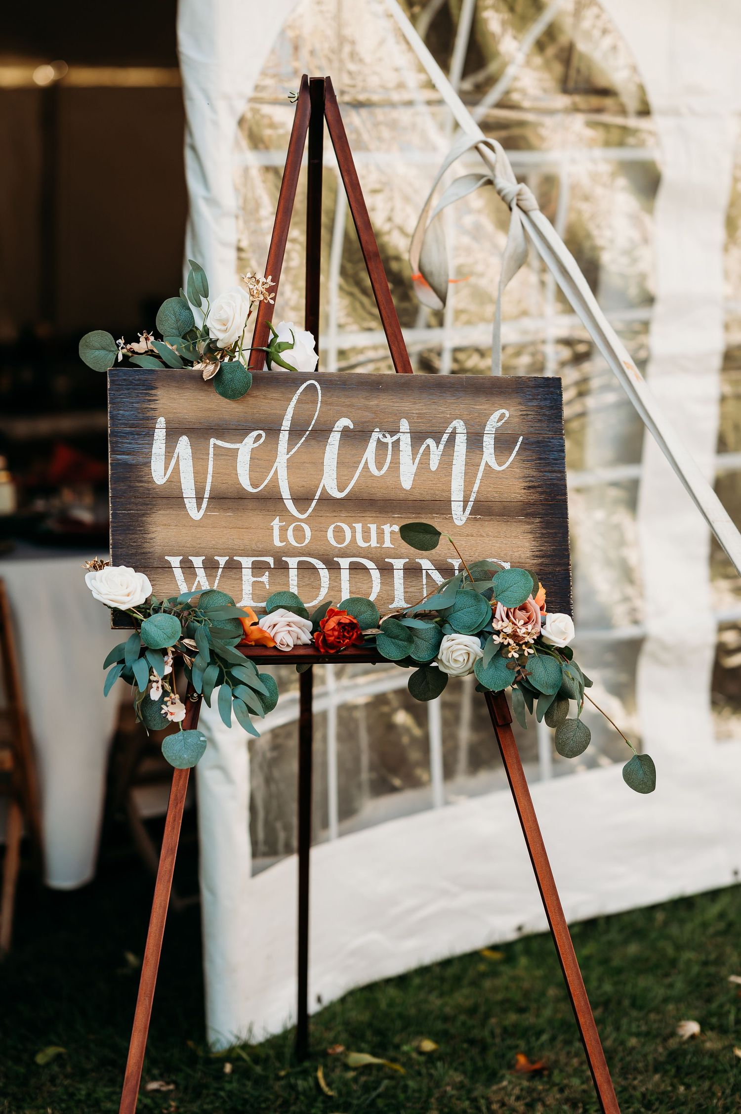 Rustic wooden welcome sign decorated with eucalyptus leaves and flowers on a wooden easel stand at an outdoor event.