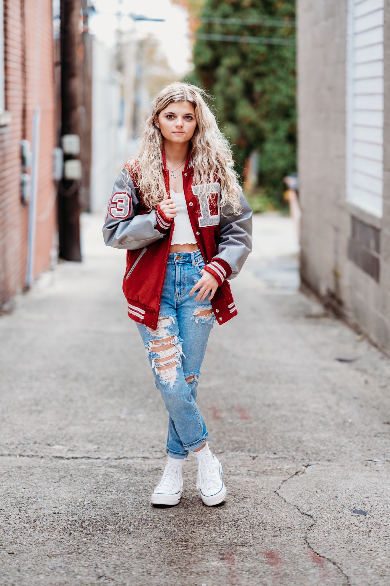 Trendy letterman jacket and distressed denim look styled with white boots in urban alleyway setting for fashion photoshoot.