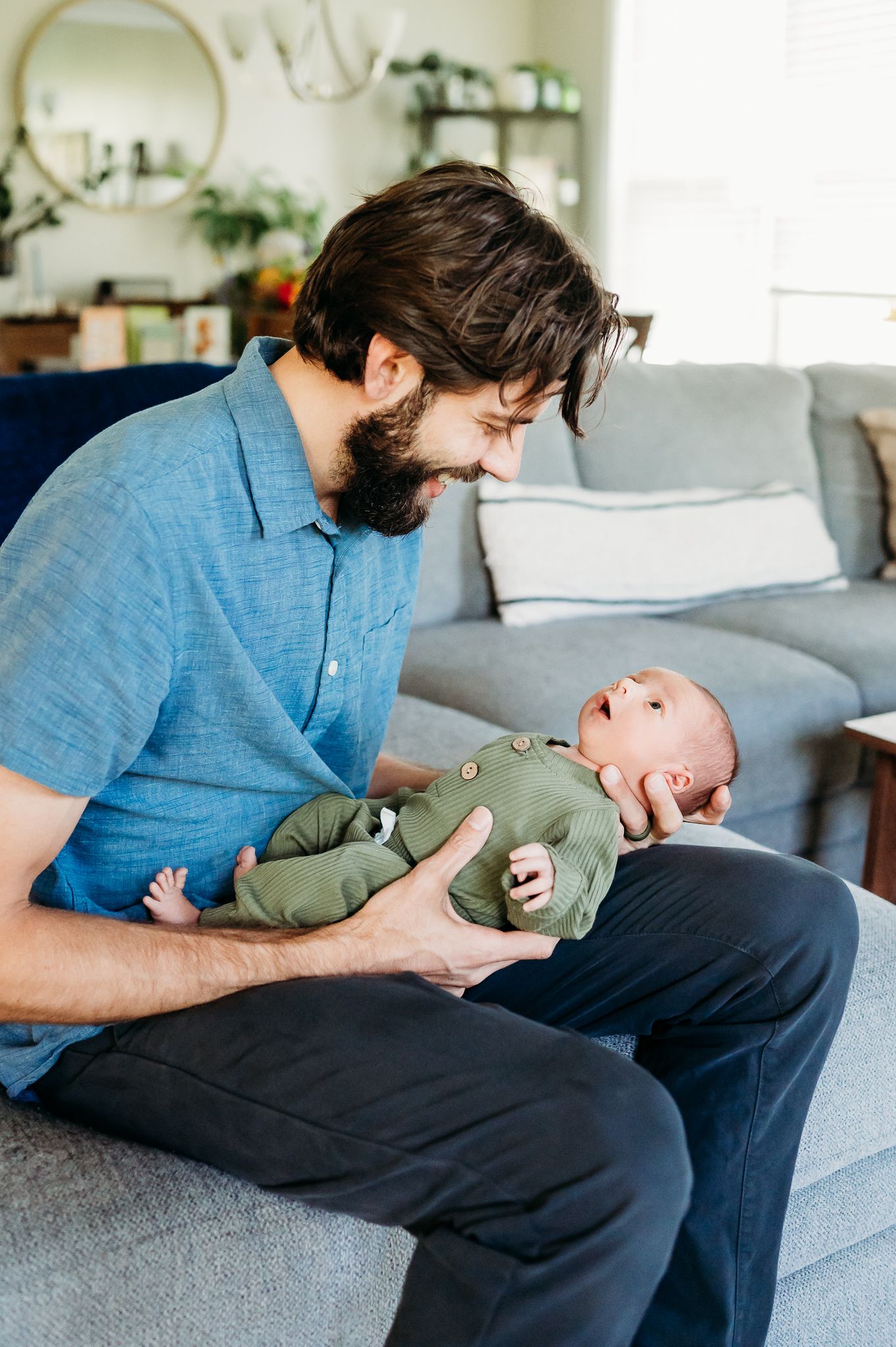 In a sunlit living room with a gray couch, a bearded man in blue shirt shares a joyful moment with a baby in green.