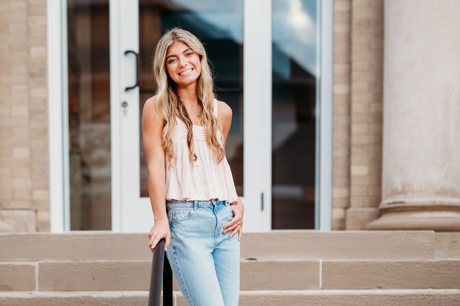 A cheerful person in a pink tank top and distressed jeans leans against a stone pillar on steps.