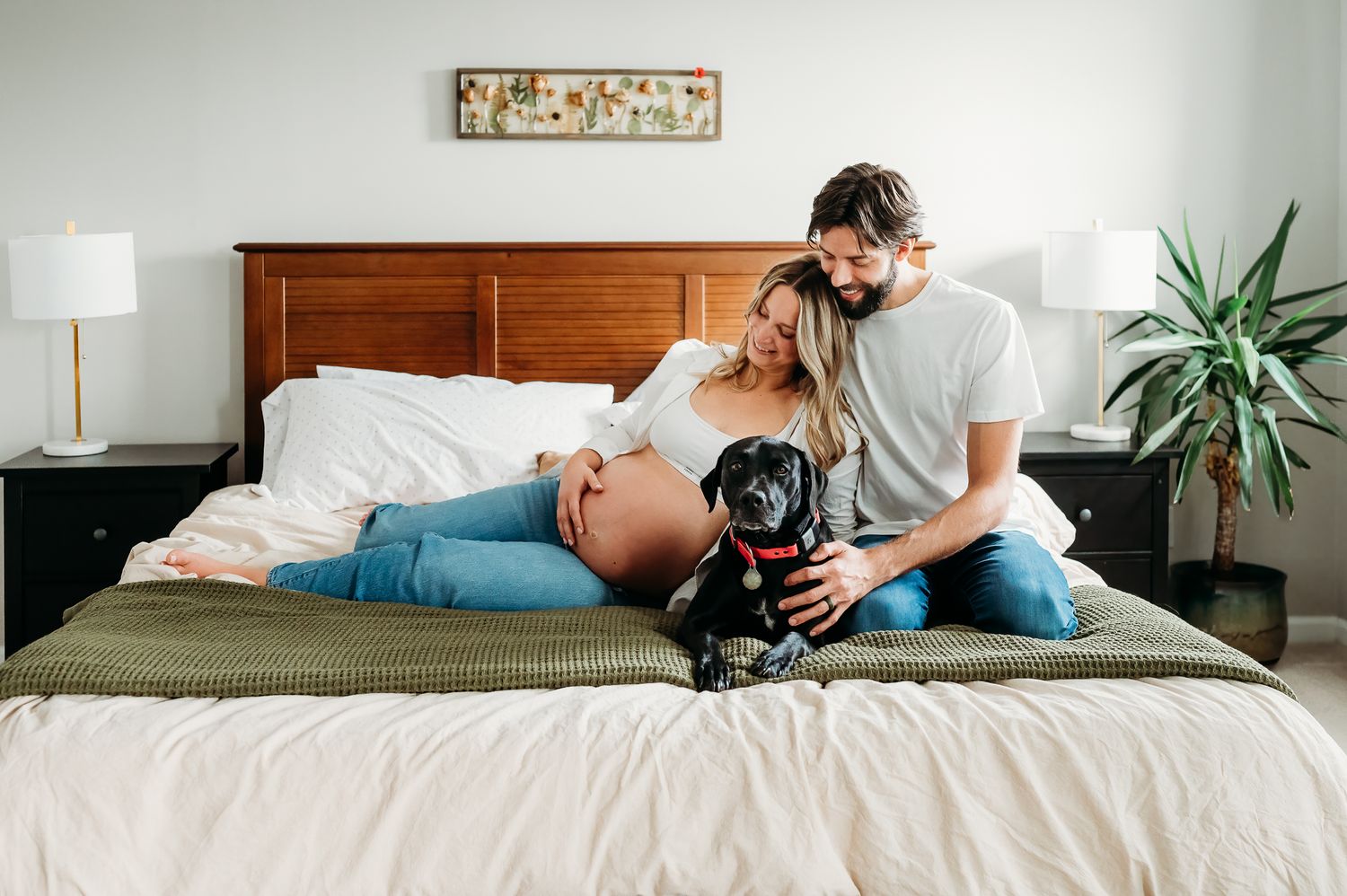 Man and pregnant woman sit together on a bed with a large black dog while relaxing in a cozy bedroom with warm decor.