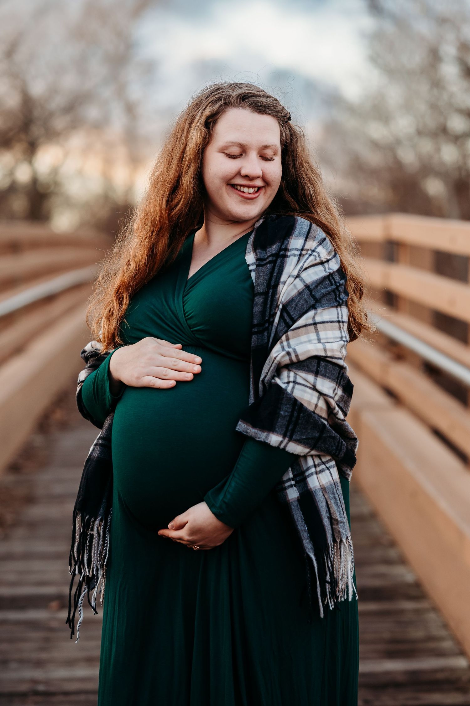 Smiling pregnant woman in green dress and plaid shawl cradling baby bump on wooden bridge outdoors.