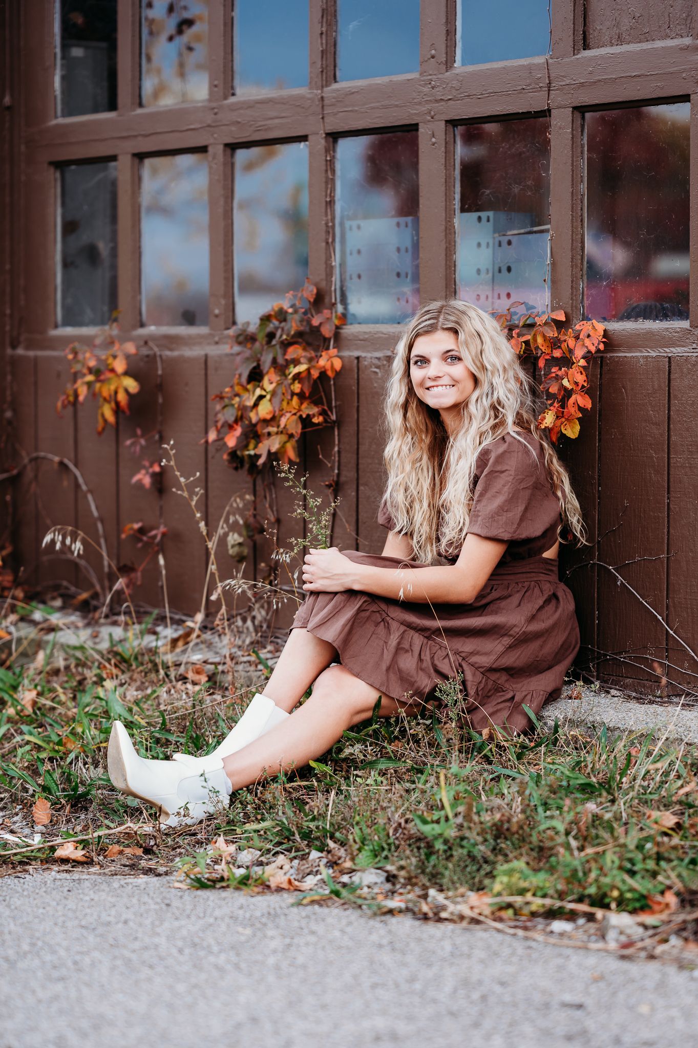 A person in a brown dress sits on the ground against a rustic door with autumn vines and white boots.