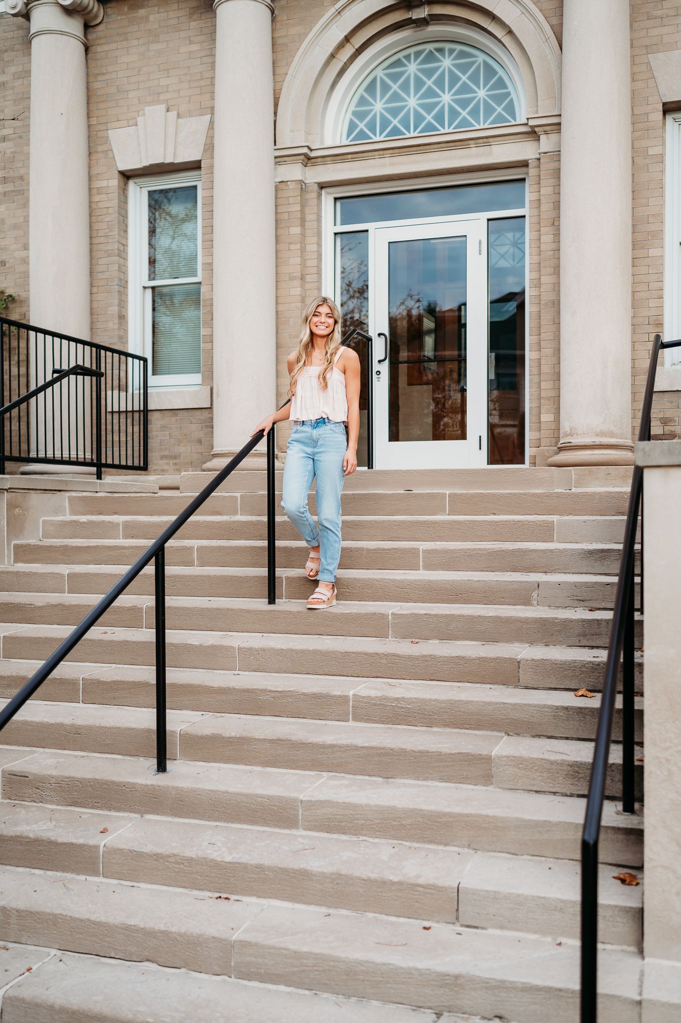 A person stands on stone steps leading to a grand entrance with columns and an arched window above glass doors.