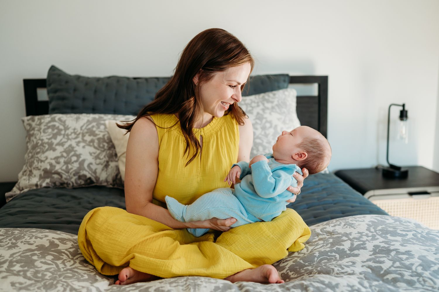 Woman in yellow dress sits cross-legged on gray bedding while holding infant in blue outfit.