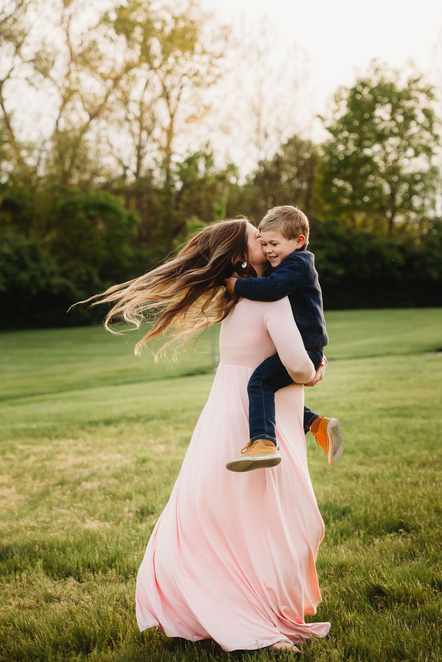 Mom in flowing pink dress spins with son in navy blue on grassy field at sunset during spring maternity session at Friendship Gardens.