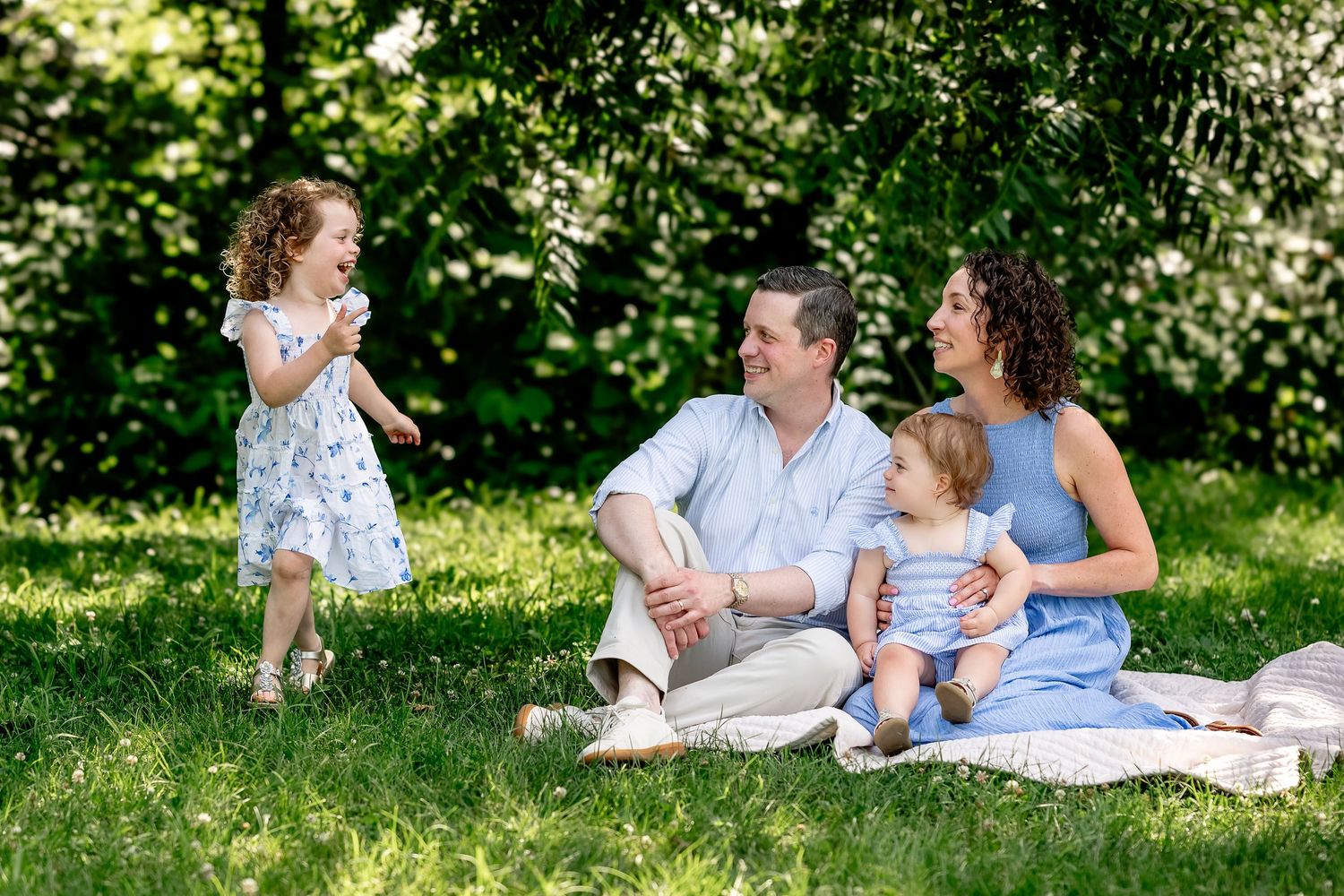 Family of four enjoying a sunny picnic on green grass, with two young children in a lush outdoor park setting.