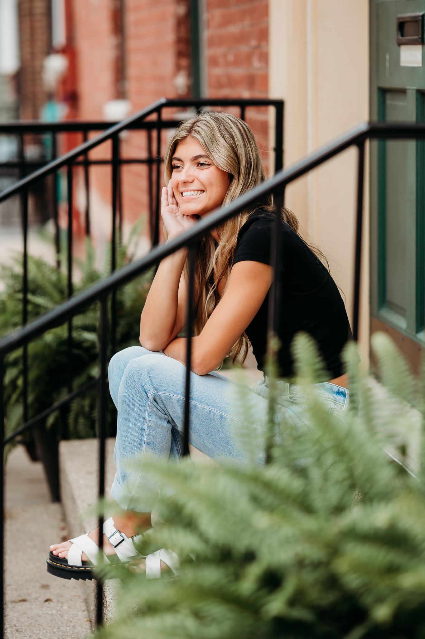 A casual outdoor portrait showing someone sitting on steps with ferns in foreground, wearing jeans and dark top.