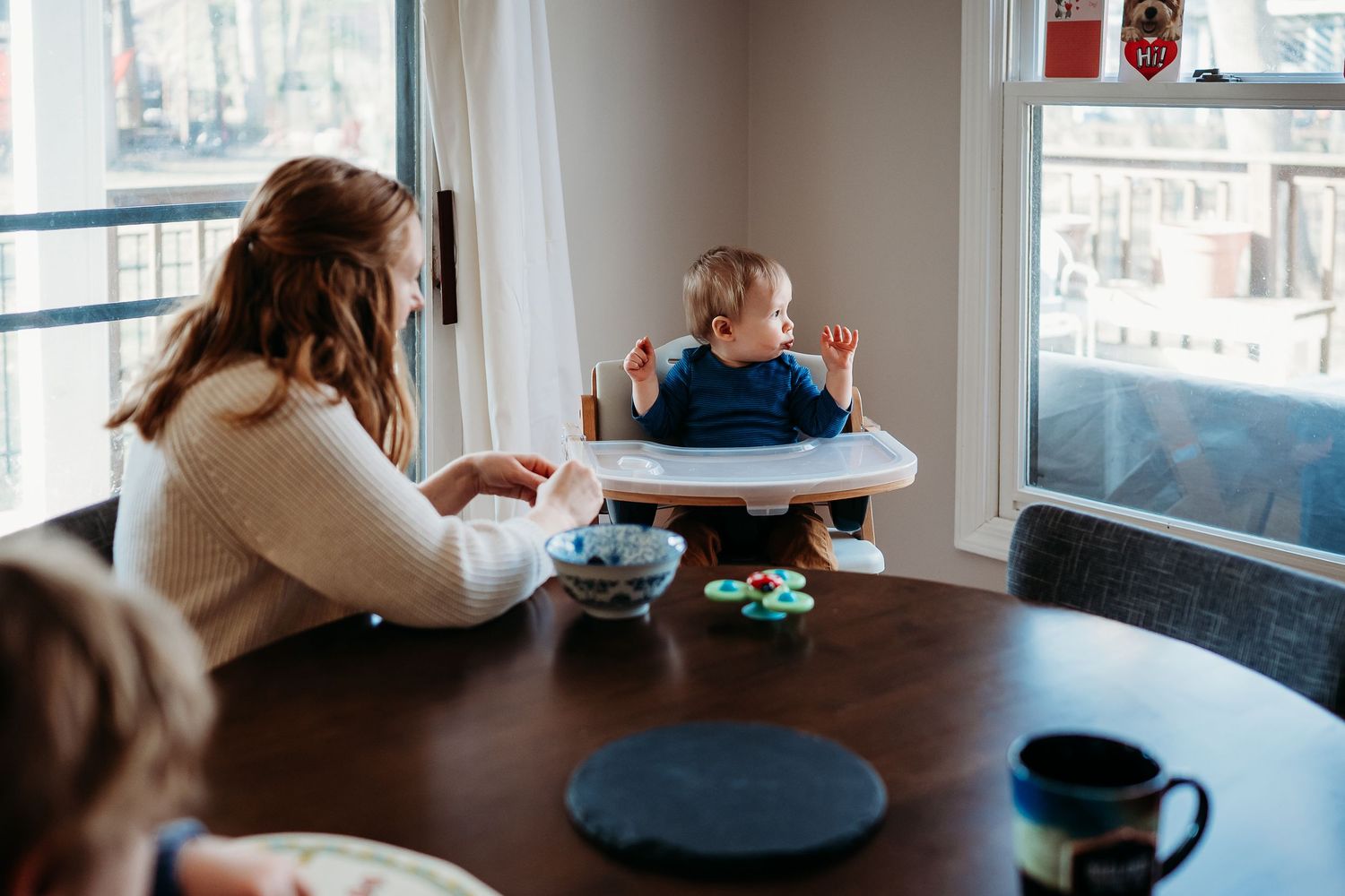 Mother feeding toddler in high chair at wooden dining table near bright windows in cozy home setting.