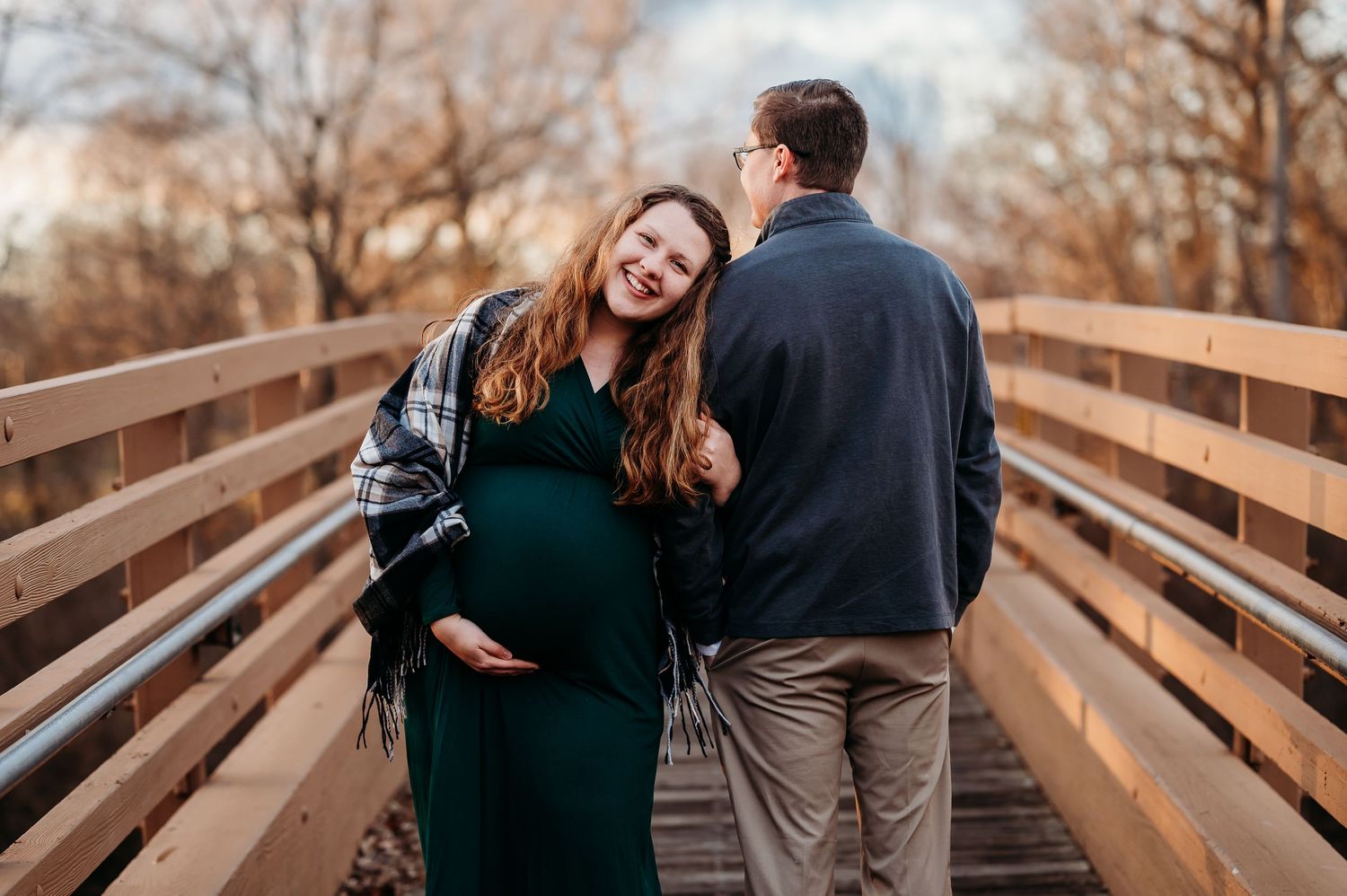Pregnant woman in green dress laughing with partner on wooden bridge during maternity photo session in Plainfield, Indiana.