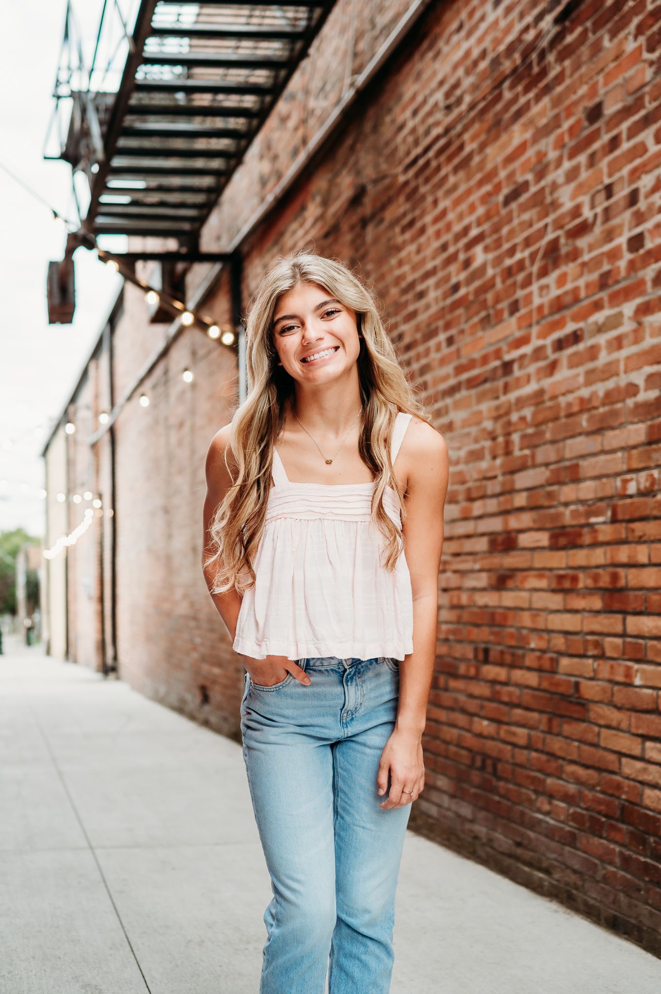 Young woman in a white sleeveless top and light jeans smiles while standing against a brick wall on a city sidewalk.