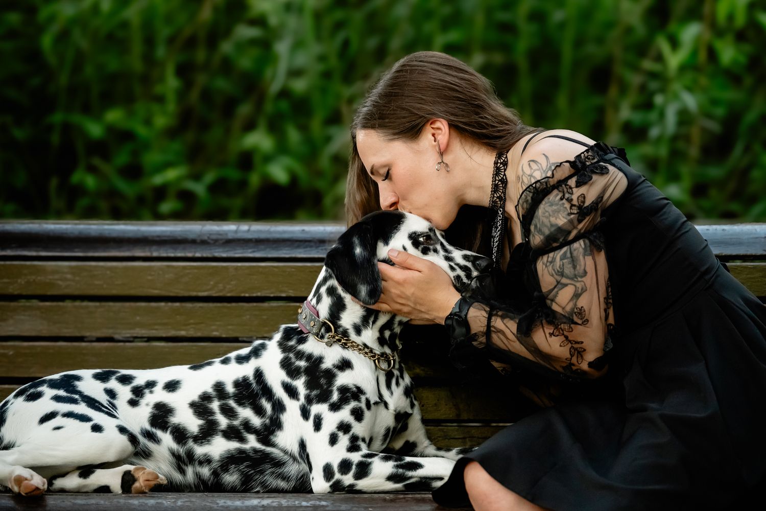 A woman in a black lace dress kisses her Dalmatian dog while sitting on a park bench.
