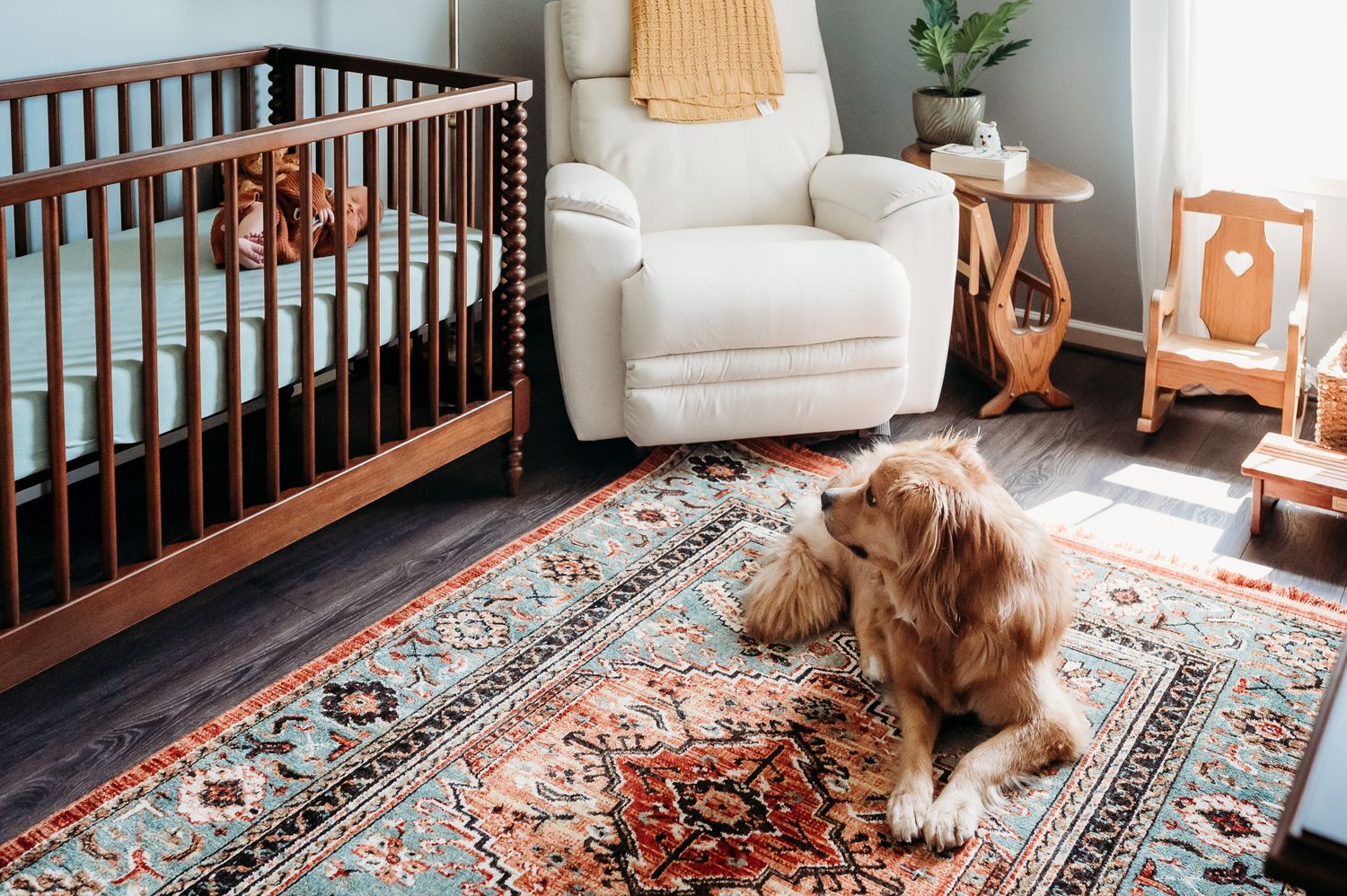 A brown and white dog lies on colorful patterned area rug beside a wooden crib in a nursery during Indiana in-home newborn photo session.