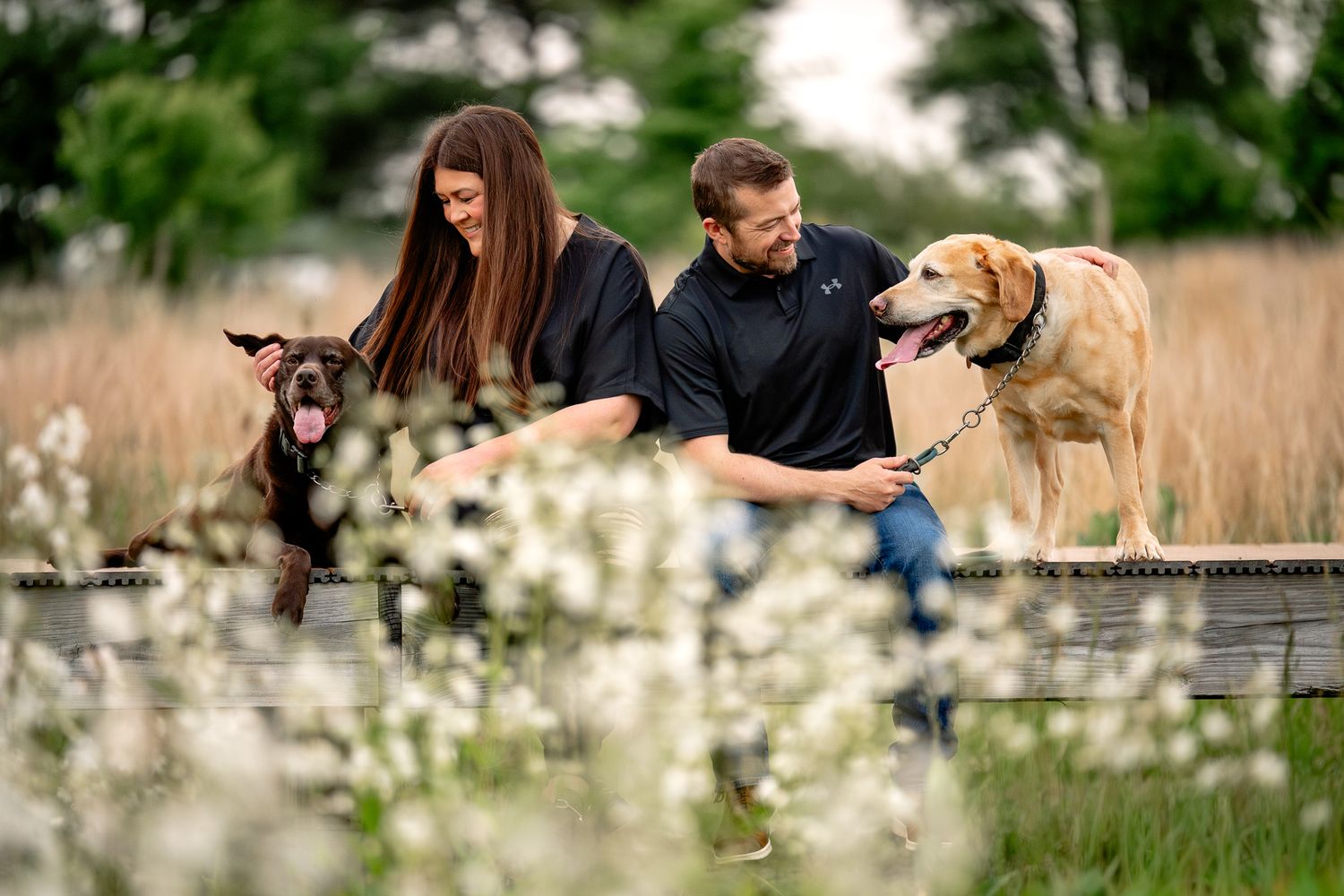 Couple in black outfits posing outdoors with two dogs on a wooden deck surrounded by blooming white flowers.