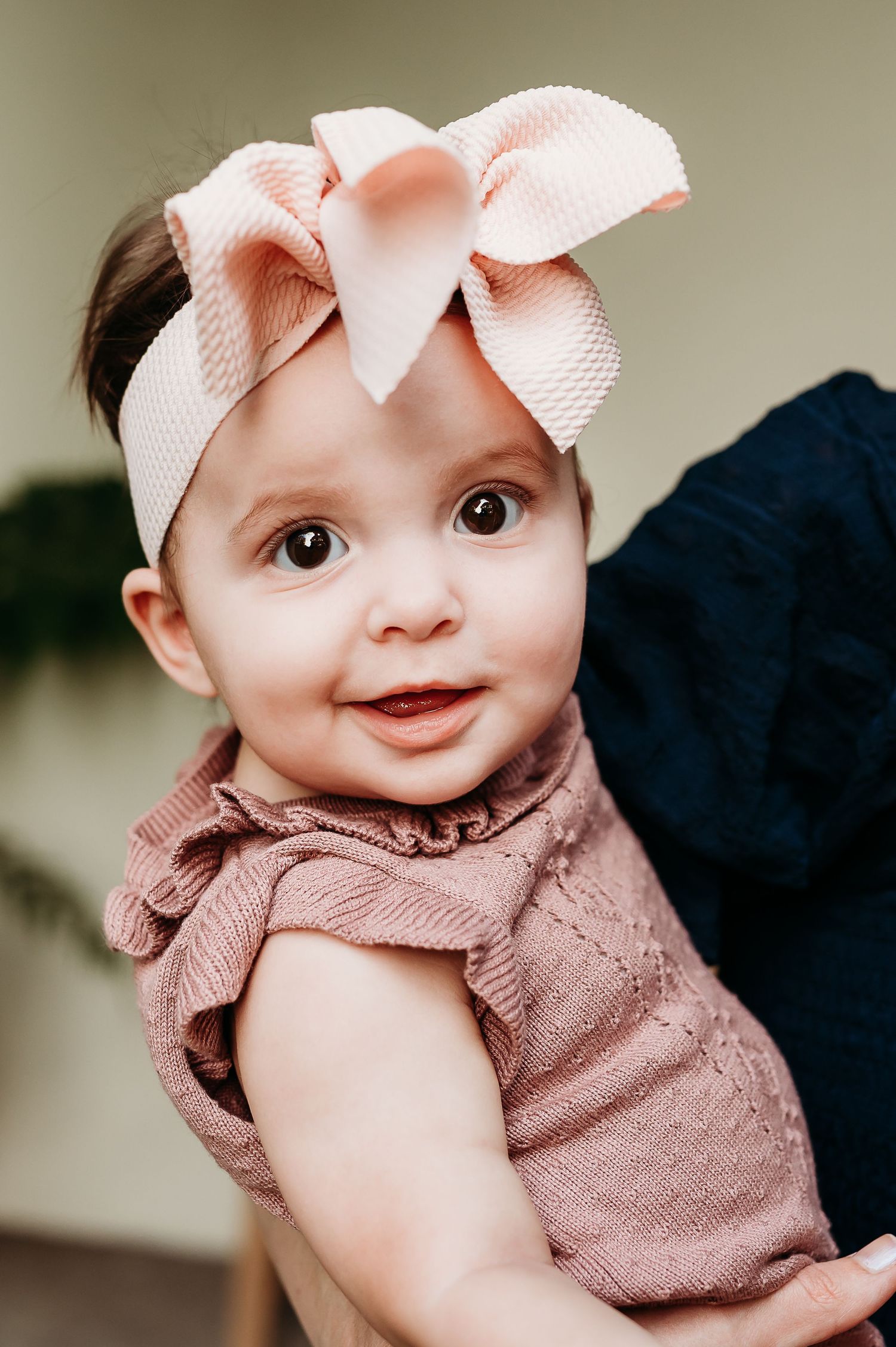Sweet baby wearing a pink bow and mauve knit outfit smiles in a close-up portrait.