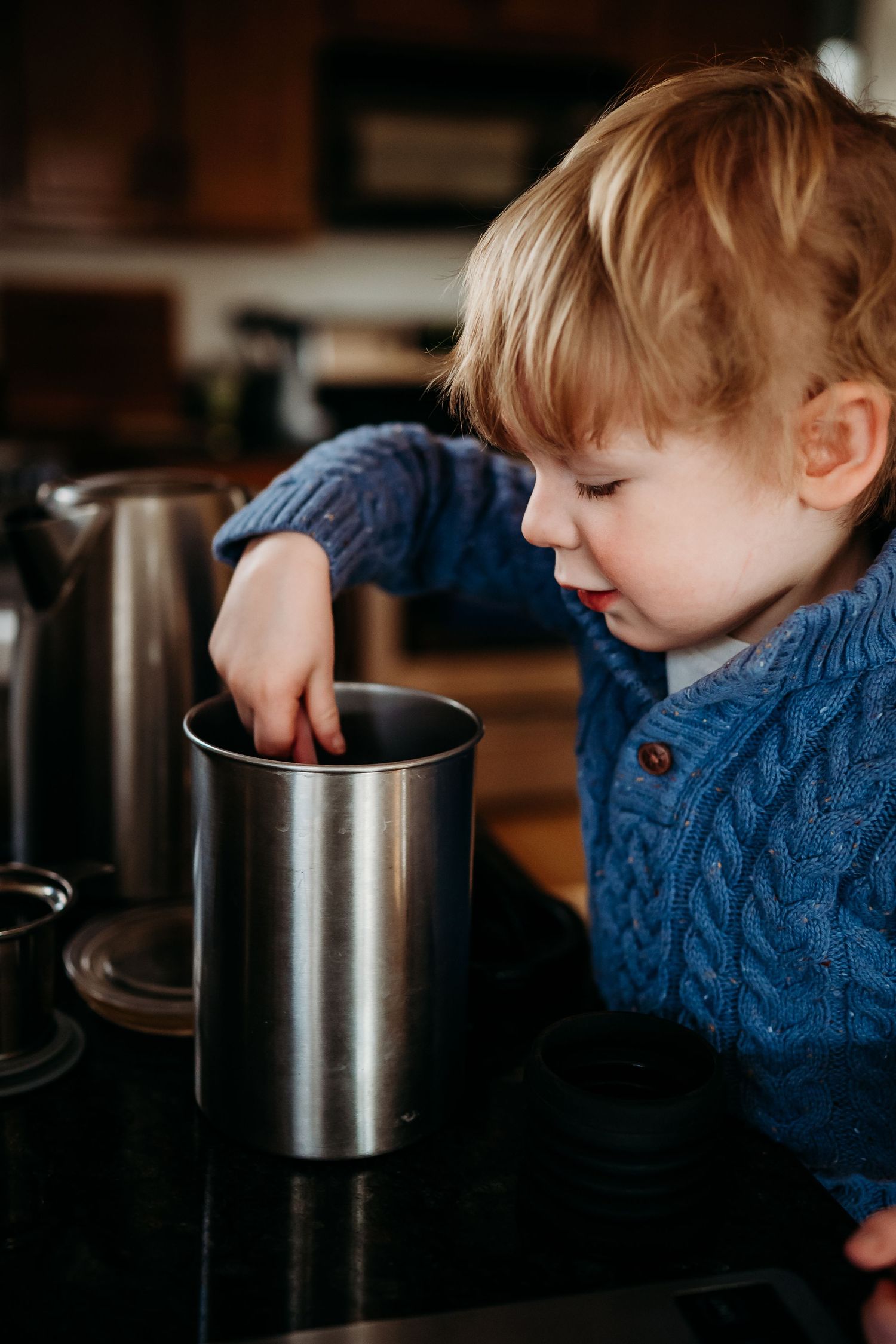 Curious toddler in blue knit sweater reaching into a stainless steel coffee cup on a kitchen counter.