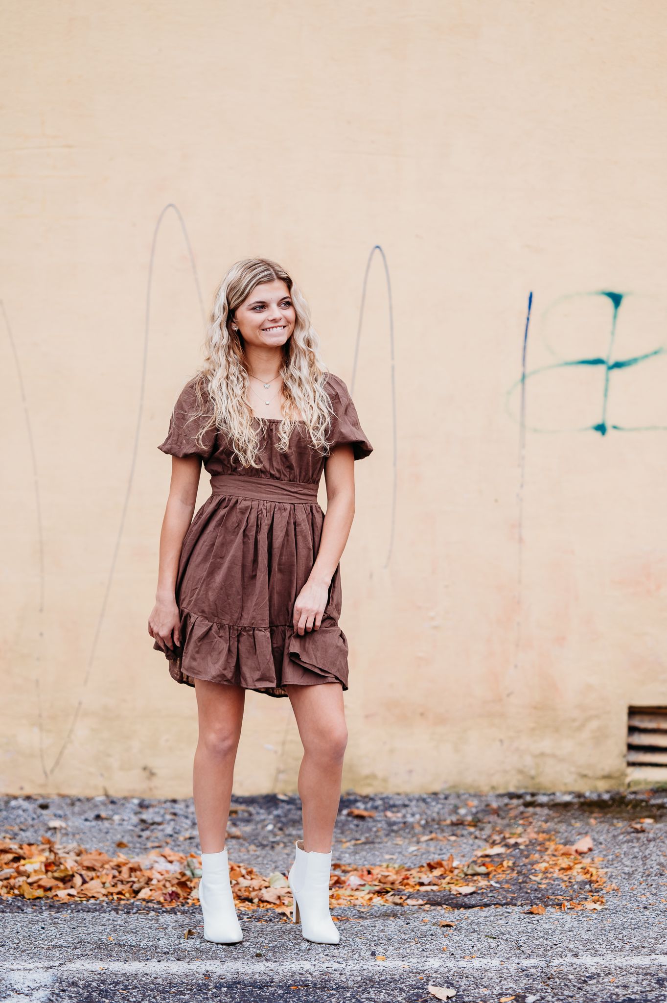 Person in a brown ruffle dress and white boots poses casually against a beige wall with autumn leaves on the ground.