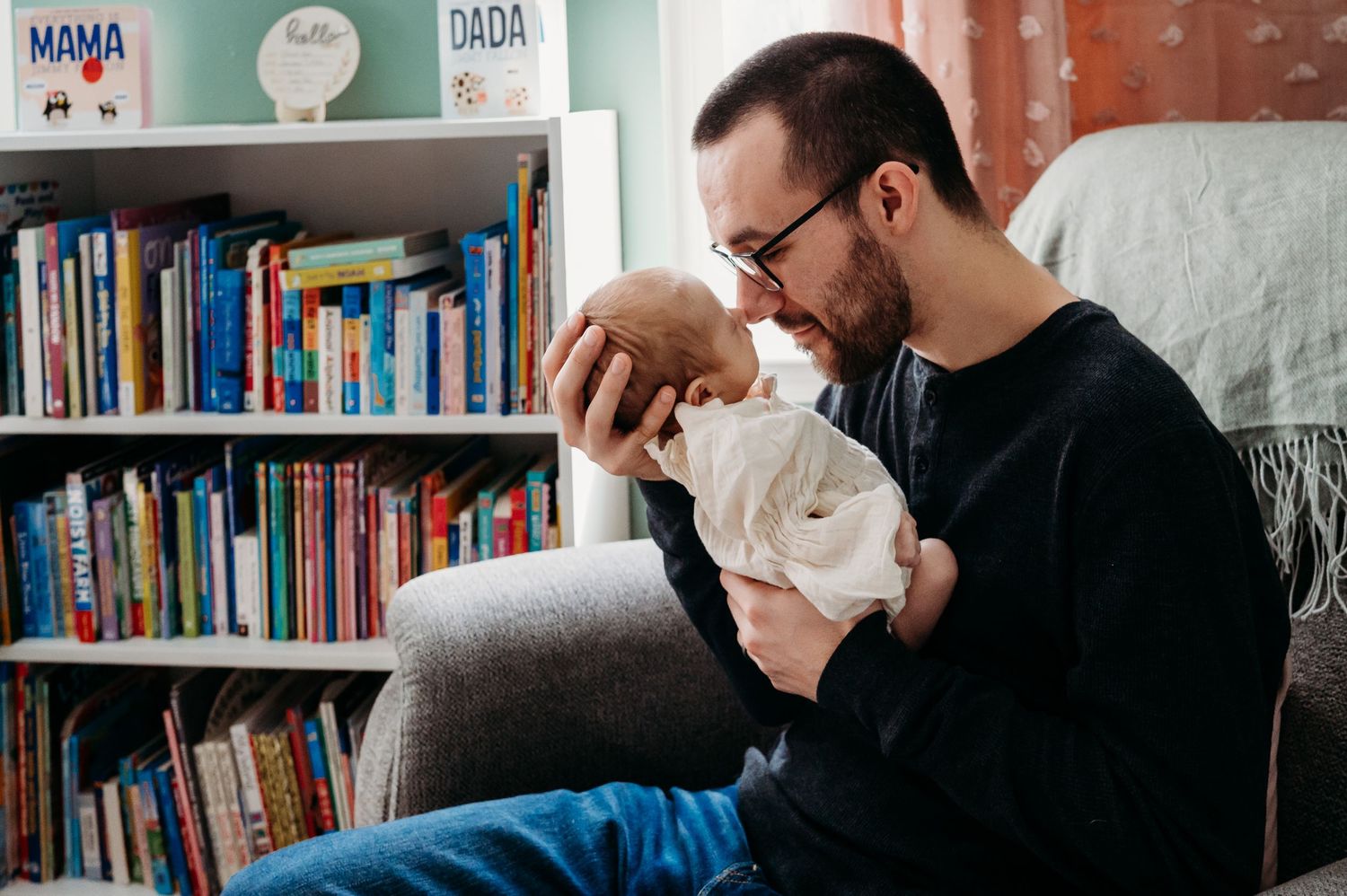 A father with glasses tenderly holds his newborn baby while sitting in a chair next to a colorful bookshelf.