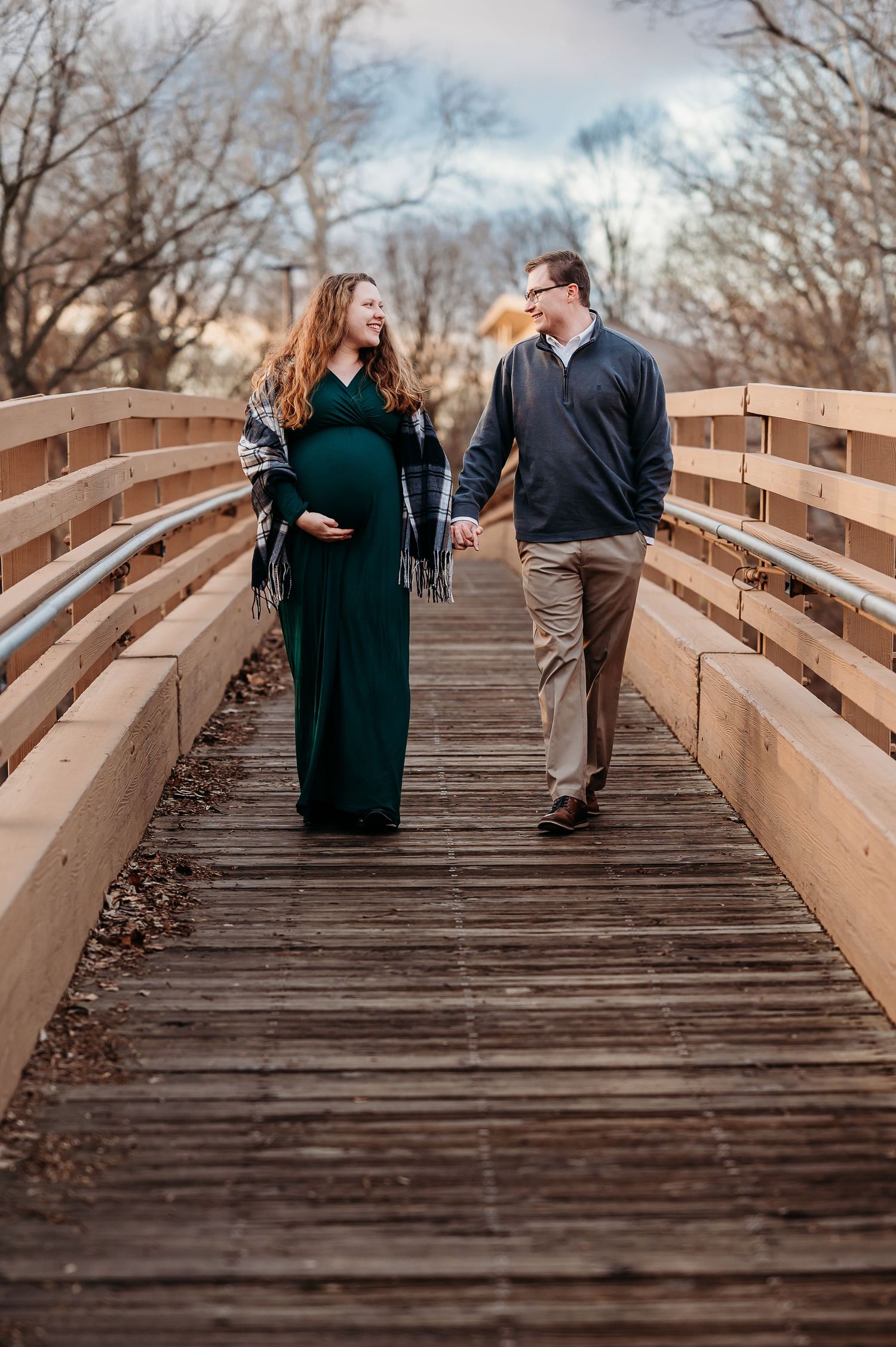 Pregnant couple holding hands walking on wooden bridge in Plainfield, Indiana, woman in green dress, during maternity photo session.