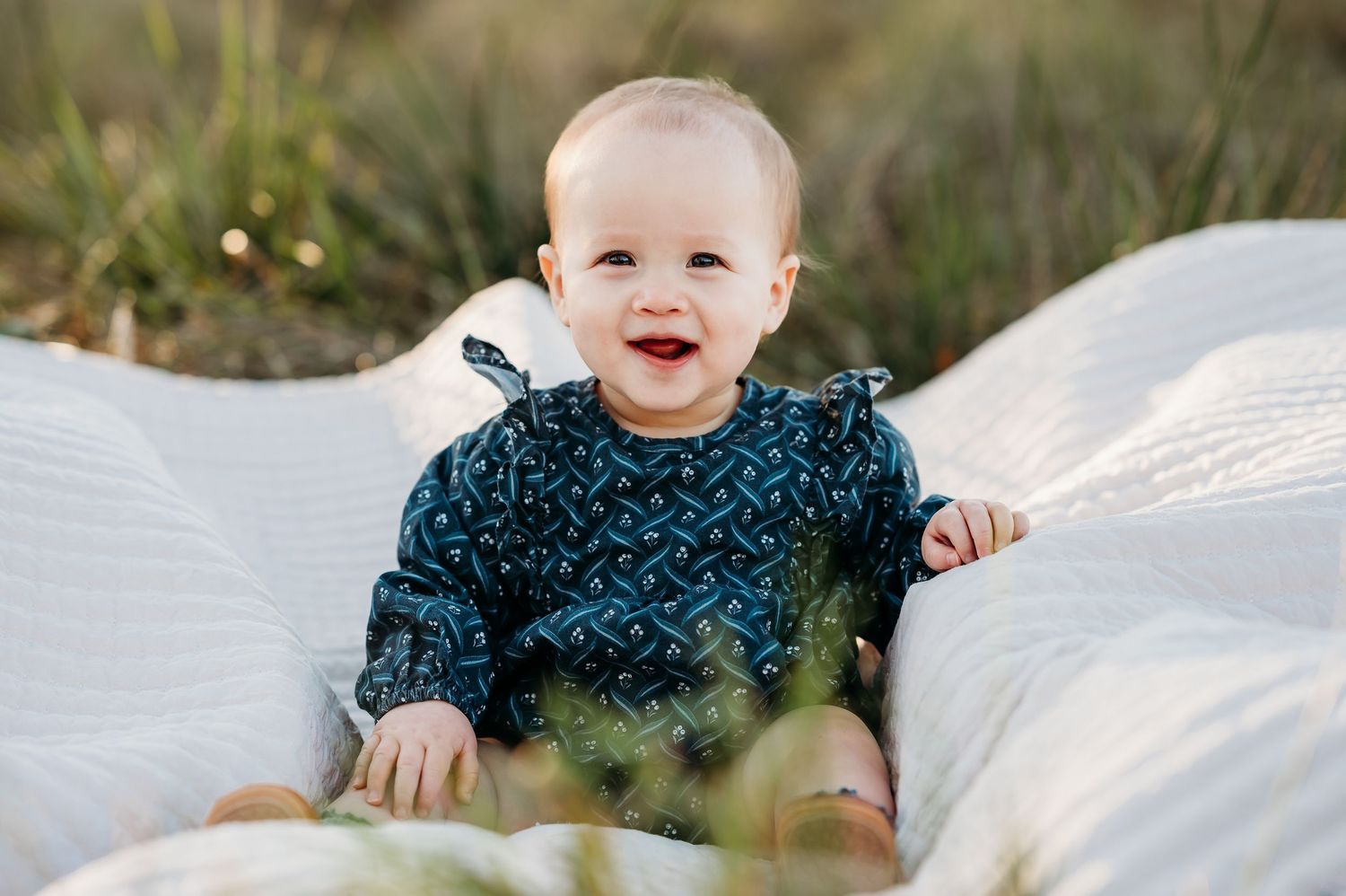 Baby wearing a blue patterned sweater sits smiling on white blankets outdoors in natural setting.