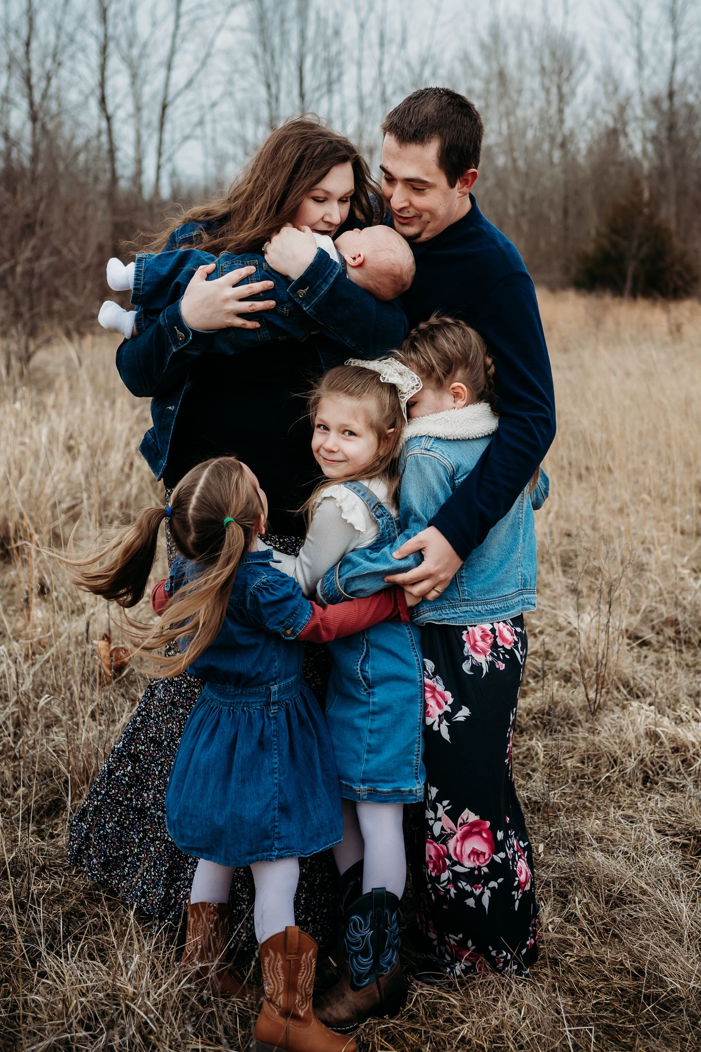 An Avon, Indiana, family embraces together in a golden field during overcast family photoshoot while wearing coordinating blue outfits.