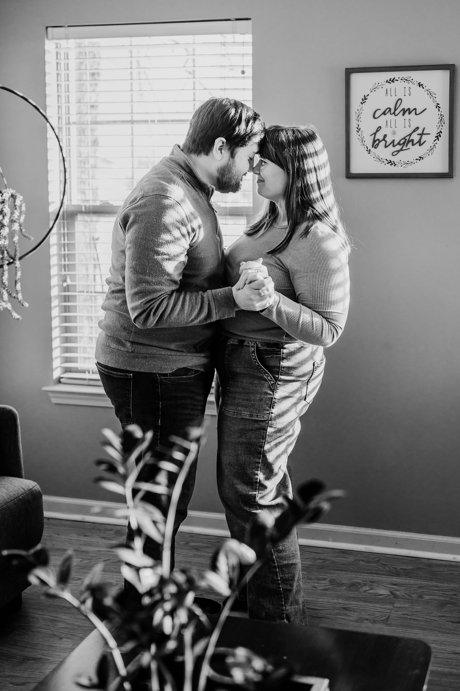 Black and white photo of a couple slow dancing and nearly kissing in a cozy home with a calm and bright sign on the wall.