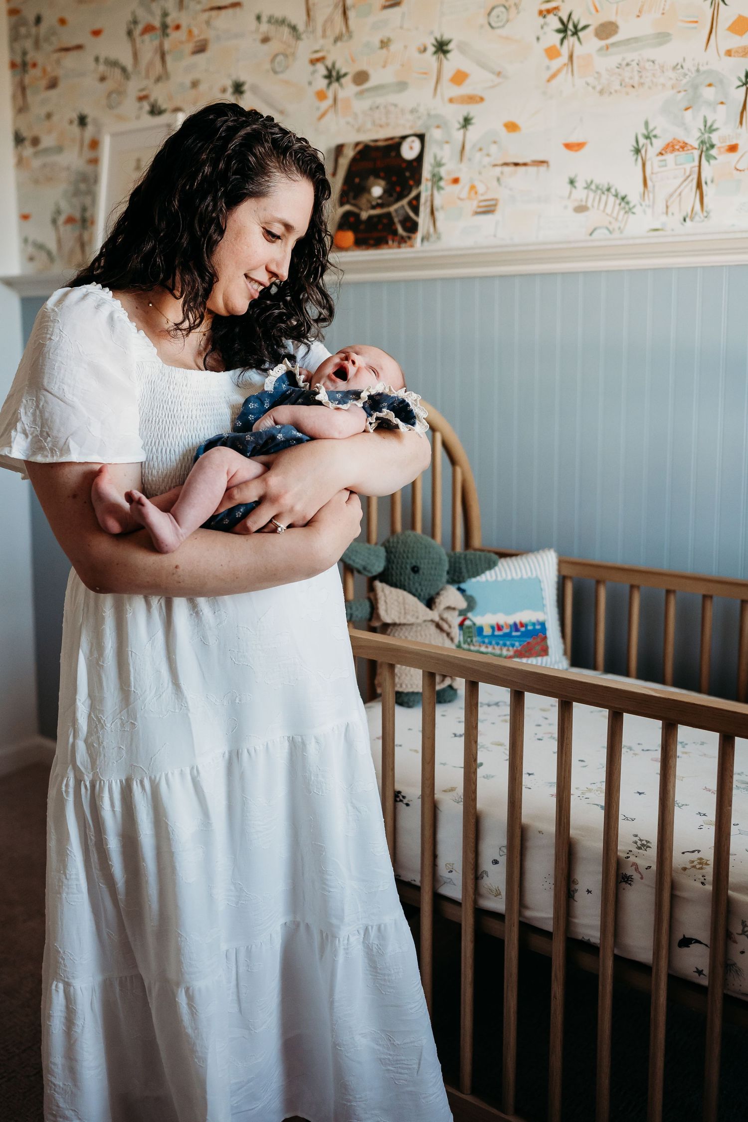 A smiling mother in a white dress holds her newborn baby beside a wooden crib in a decorated nursery.