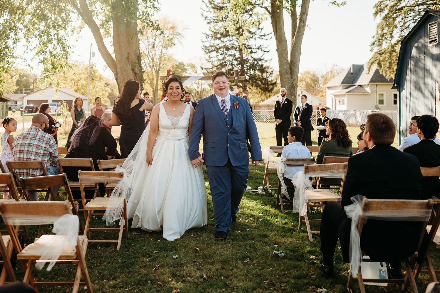 Outdoor wedding reception setup with rustic wooden picnic tables and guests mingling in golden sunlight.