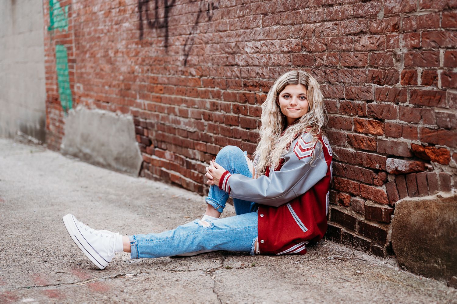 A girl in a varsity jacket and ripped jeans sits casually against a rustic brick wall in downtown Danville.