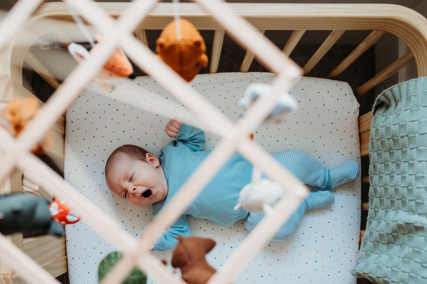 Baby in blue sleepwear rests peacefully in white crib with hanging mobile toys.