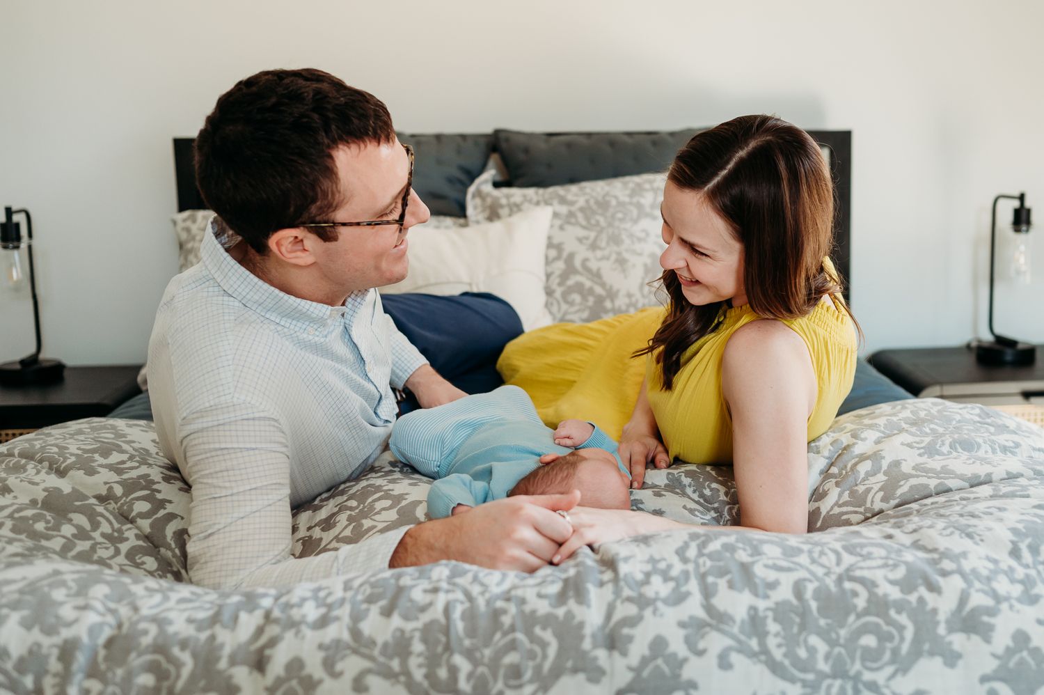 A couple relaxes together on a gray patterned bed, laughing and interacting in a bright bedroom setting with their newborn in Westfield.