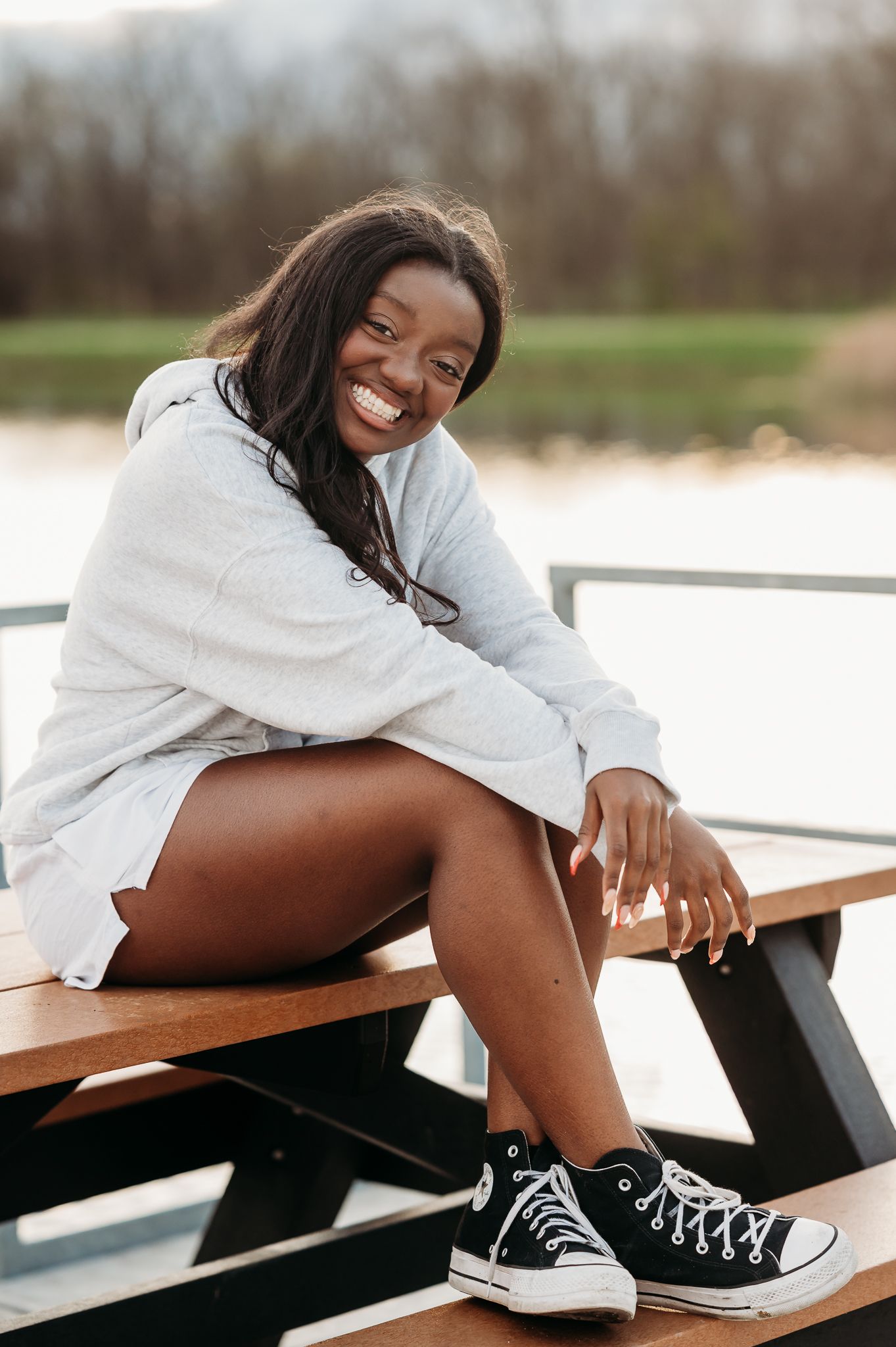 Senior girl in a white sweater and black Converse sneakers sits casually on a wooden picnic bench on a sunny day.