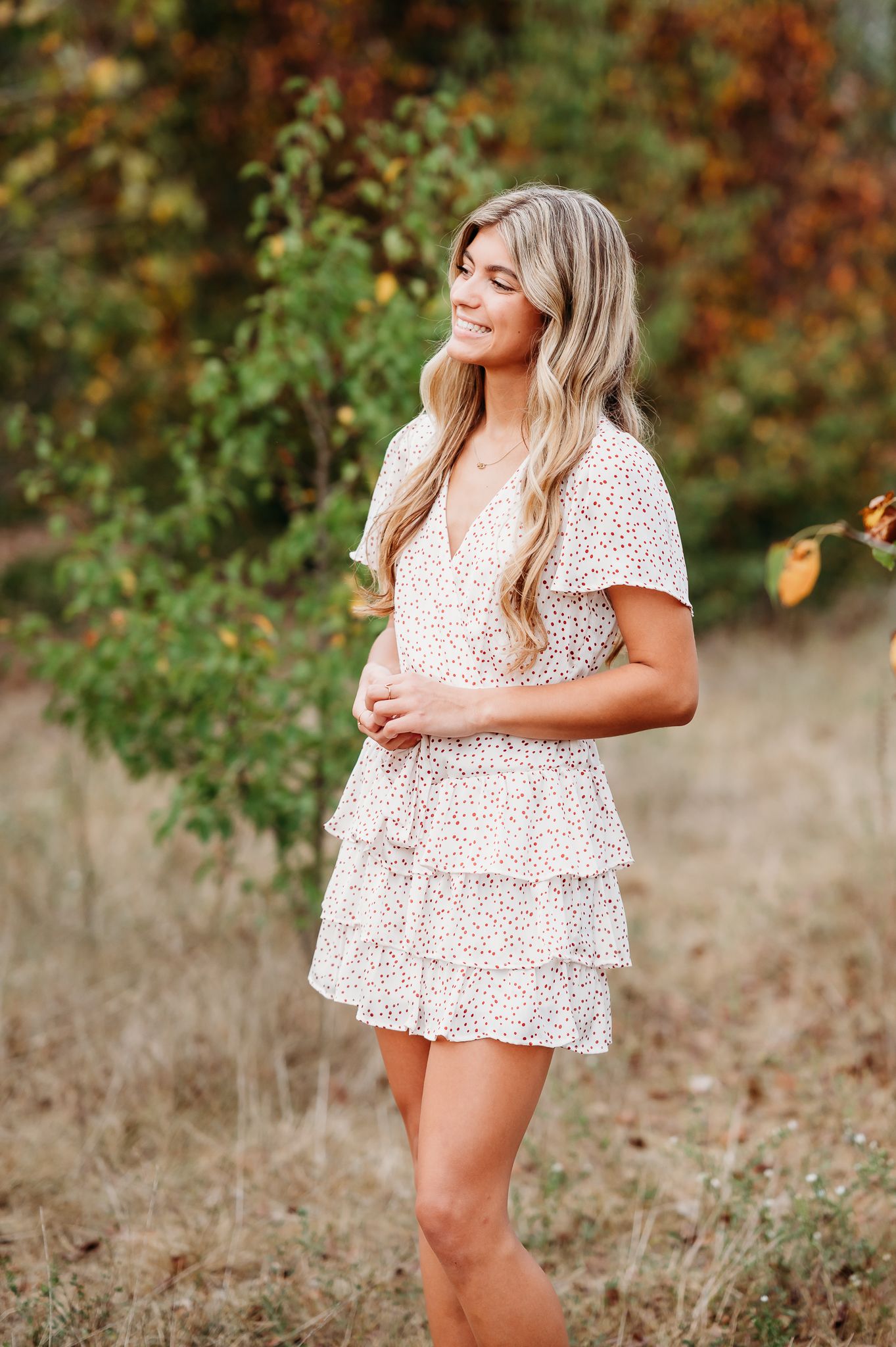 A woman in a white floral tiered mini dress poses outdoors against an autumn background.
