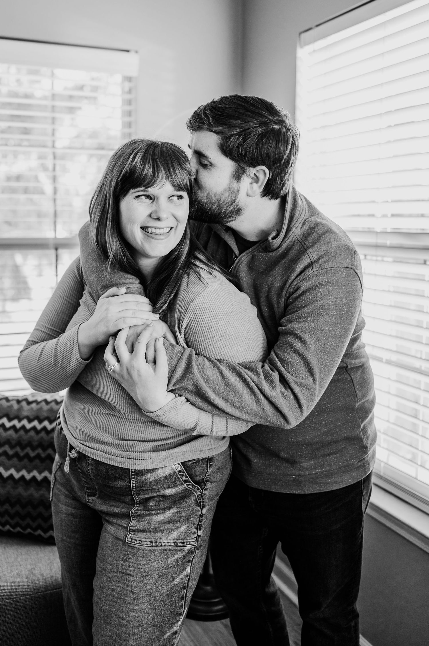 Black and white photo of a smiling woman being hugged and kissed on the cheek by a man near bright windows in Indianapolis.