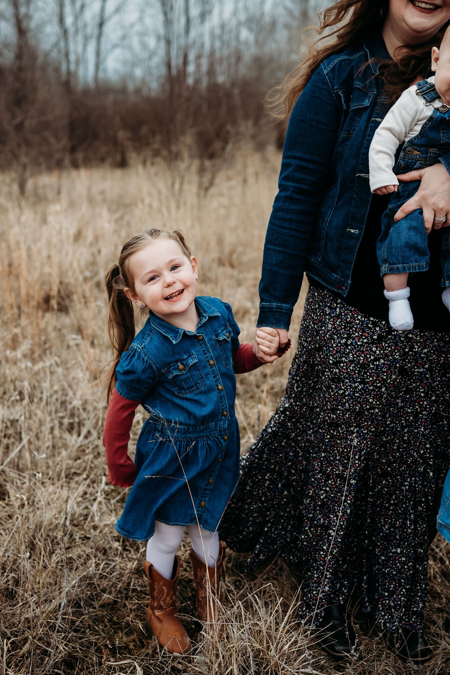 A smiling little girl in a denim dress and cowboy boots holds her mother's hand in a field.