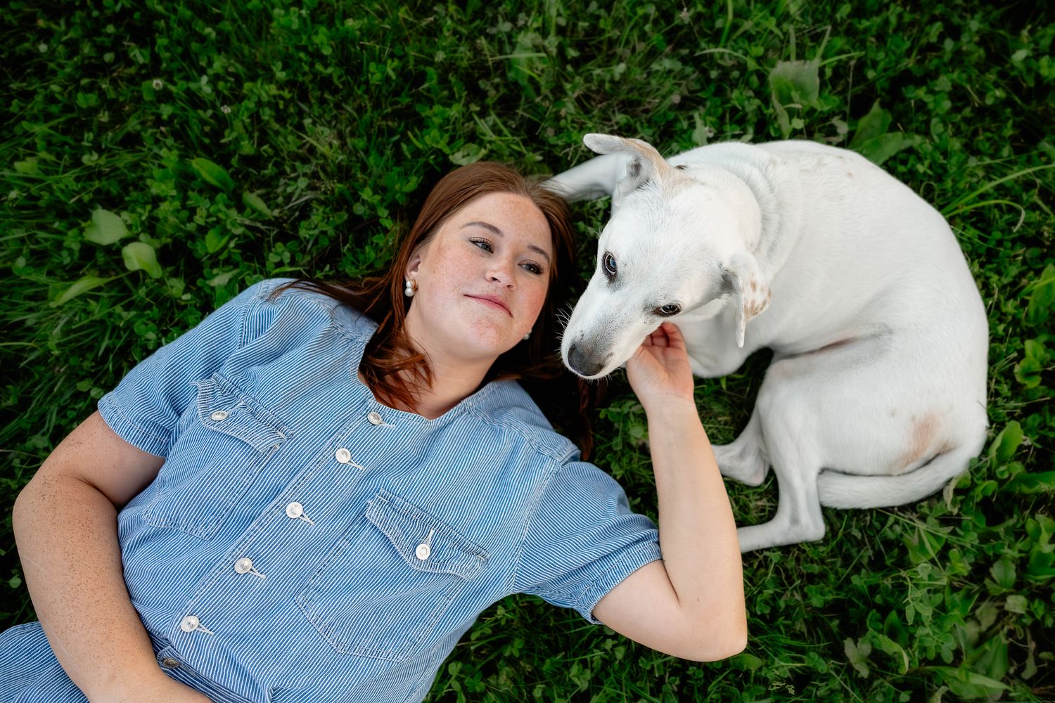 Young woman lying on green grass smiling with white dog nuzzling her face outdoors.