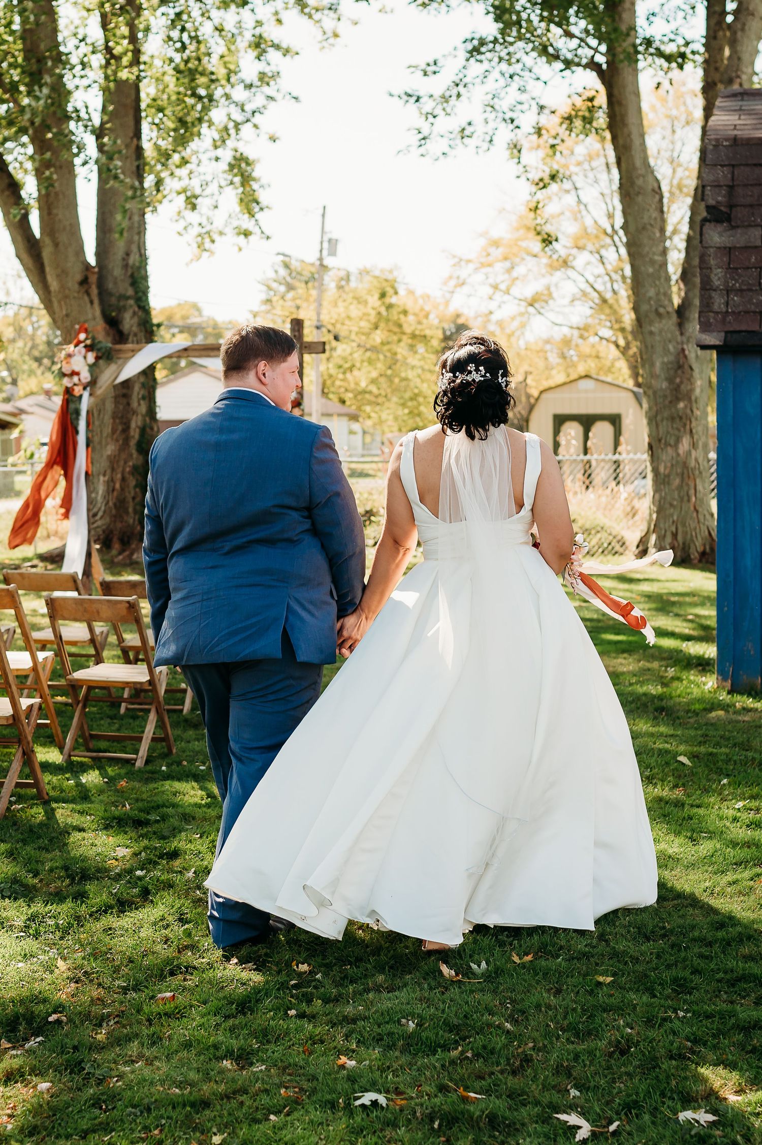 A couple in wedding attire walking together on a grassy lawn with wooden chairs and trees in the background.