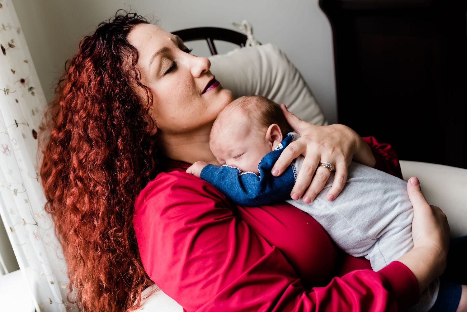 Mom with red curly hair resting with sleeping infant in blue outfit in Avon, Indiana, home nursery.