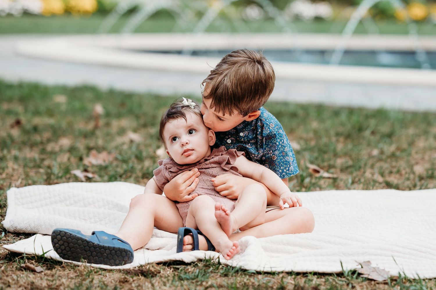 Brother gives baby sister a kiss on cheek as he holds her in front of fountain at Newfields in Indianapolis.