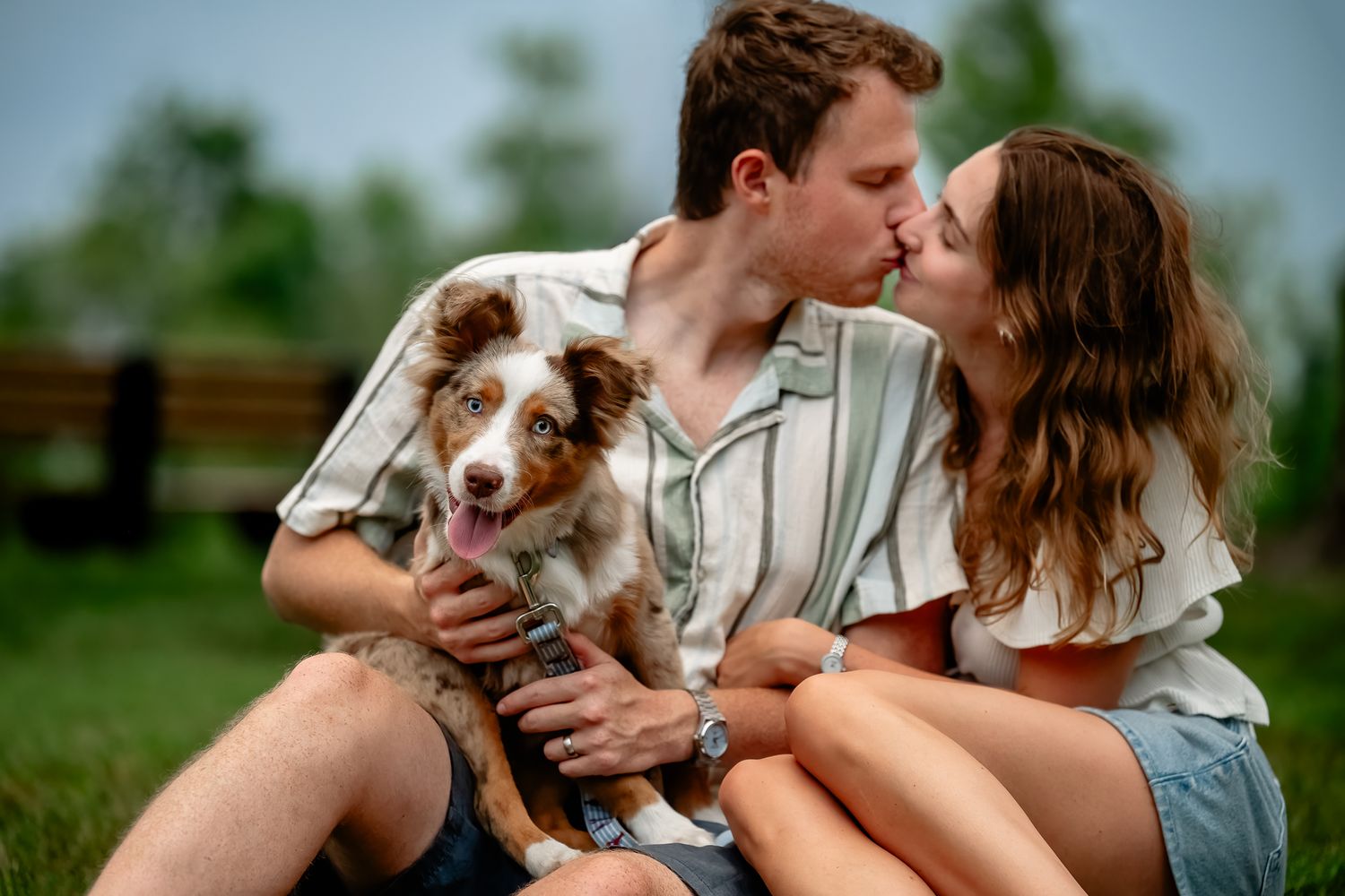 Couple kissing while holding their Mini Australian Shepherd puppy outdoors during Plainfield dog photo session with Sara House Photography.